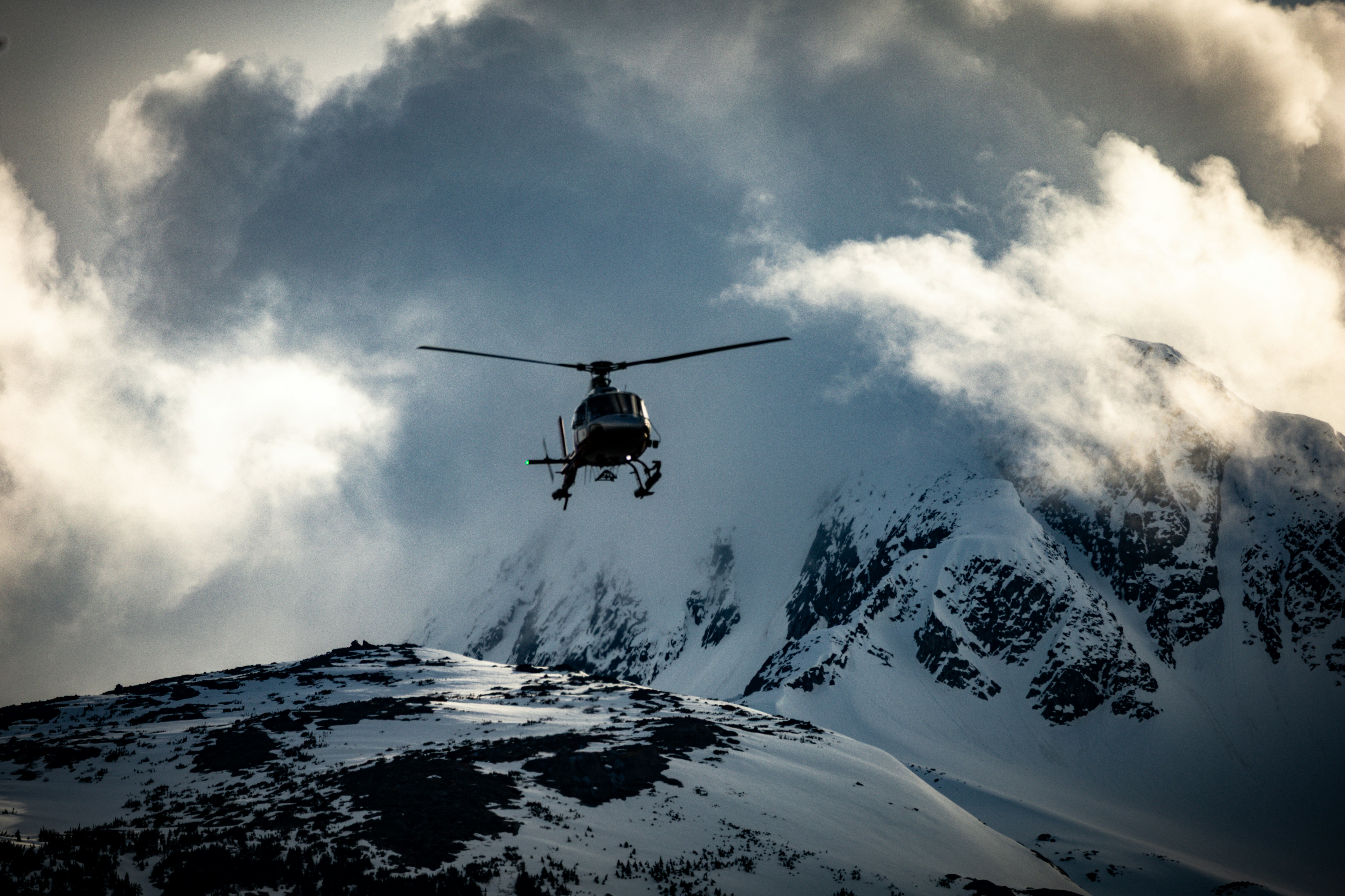 Helicopter flies over snowy mountains under dramatic clouds