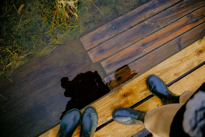 Several pairs of dark rubber boots on a wet wooden boardwalk beside marsh grass in low sun, seen from above