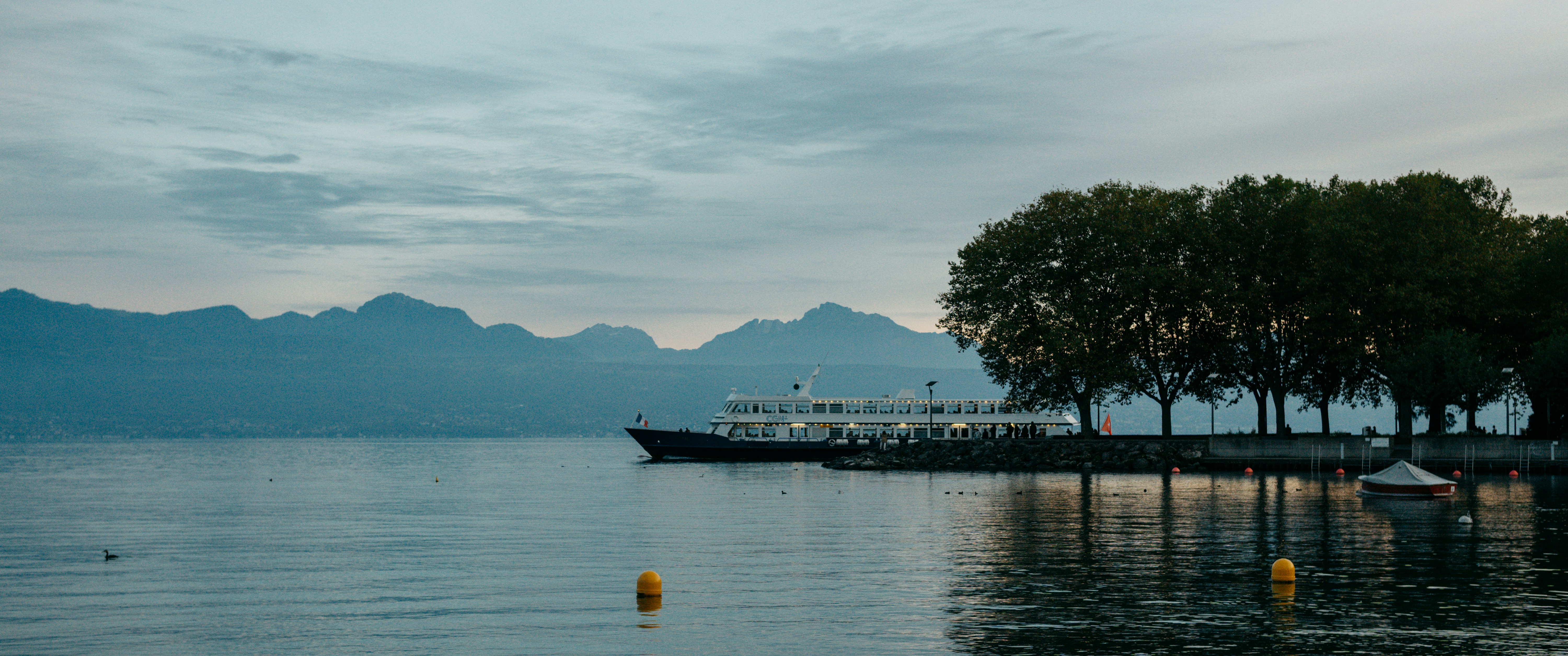 Boat docked by trees on a calm lake with mountains.