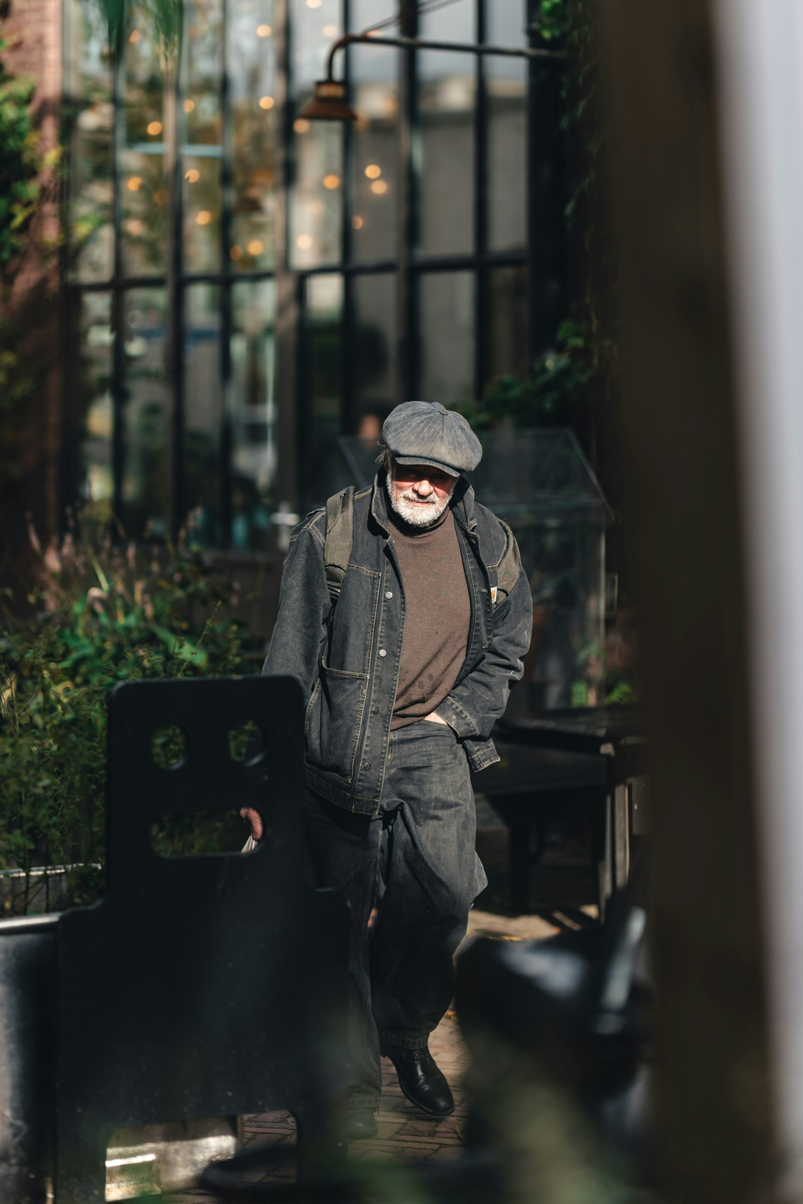 An older man wearing a cap and denim jacket walks.