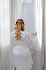 Bride in silk robe admires wedding dress hanging near window.