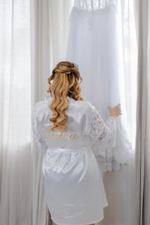 Bride in silk robe admires wedding dress hanging near window.