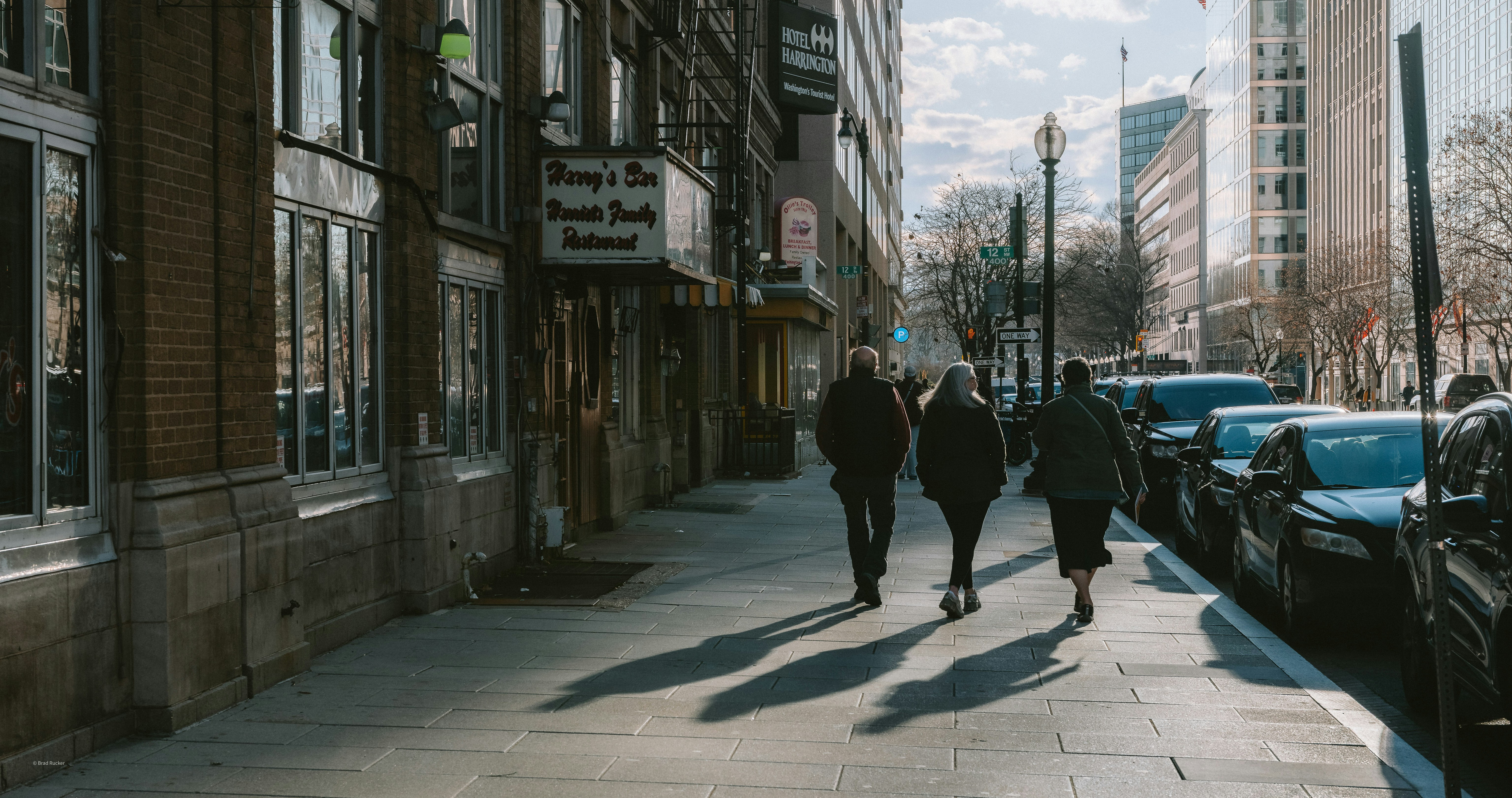 People walking on a city sidewalk next to parked cars.