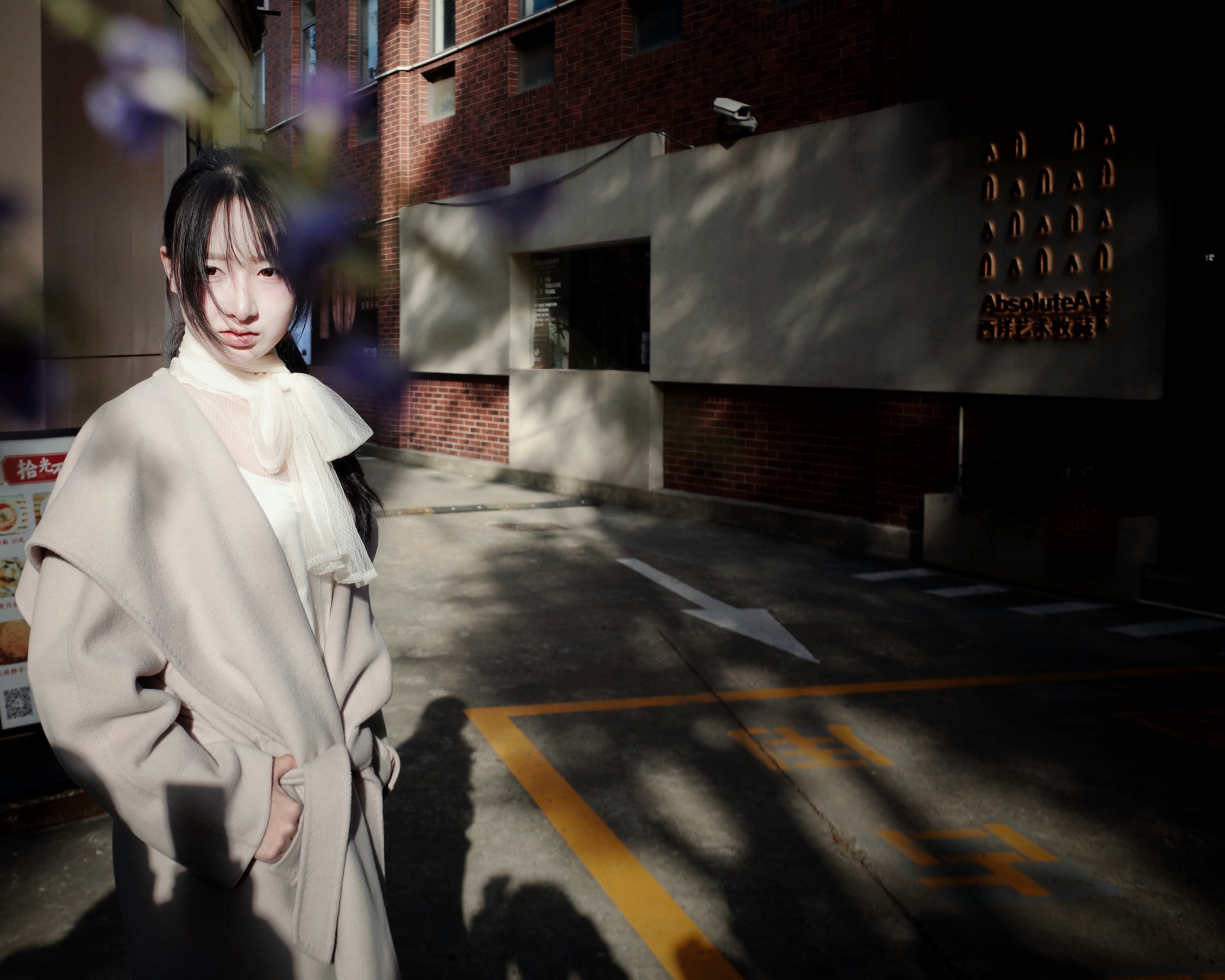 Young woman in light coat stands outdoors near brick building.