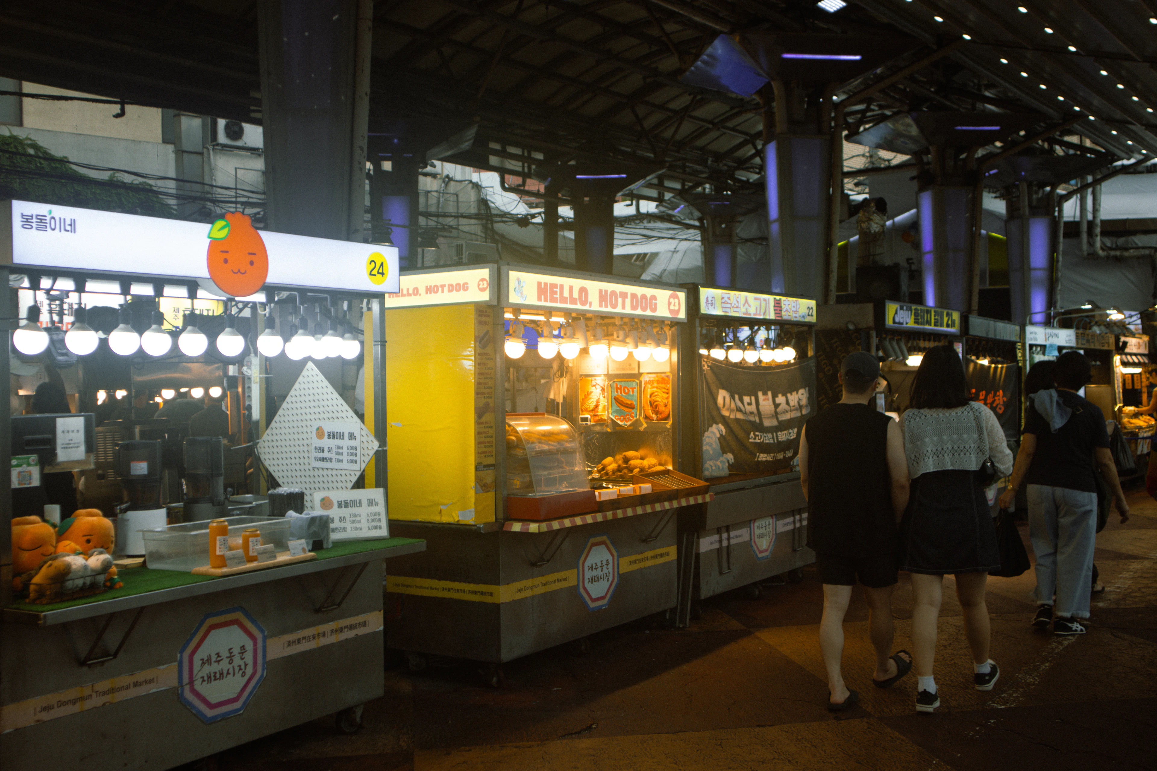 Lit food stalls at Dongmun Market in Jeju City at night, their yellow counters glowing under the covered market roof as shoppers walk the aisle between them.
