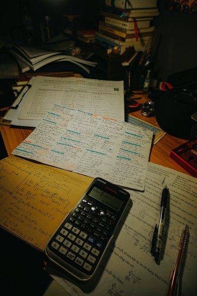 A desk covered in exam papers, a calculator, and pens under warm light