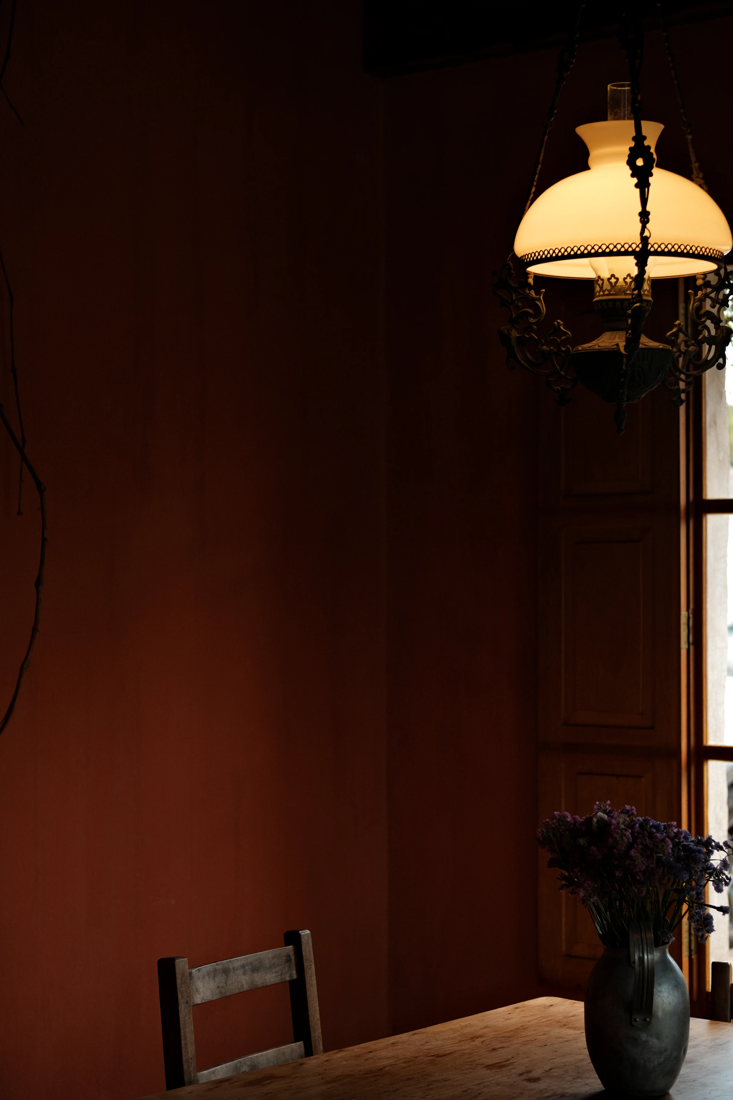 A dim living room with a wooden chair, a warm pendant lamp and dried flowers against a dark red-brown wall