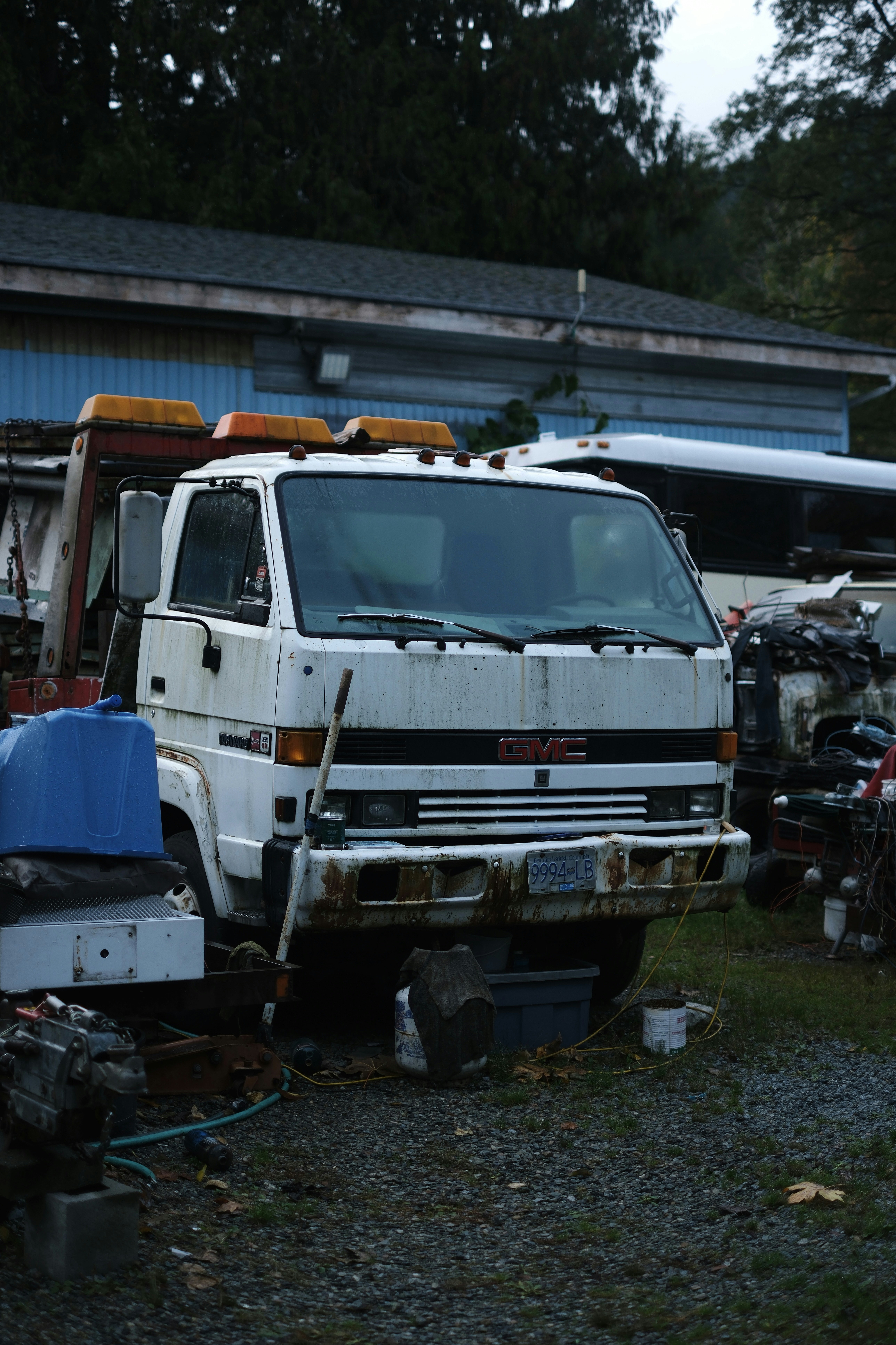 A rusty white gmc tow truck with faded paint and license plate is parked in a cluttered, overgrown lot filled with car parts, debris, and other vehicles—a reminder of the need for reliable tow truck leasing options.