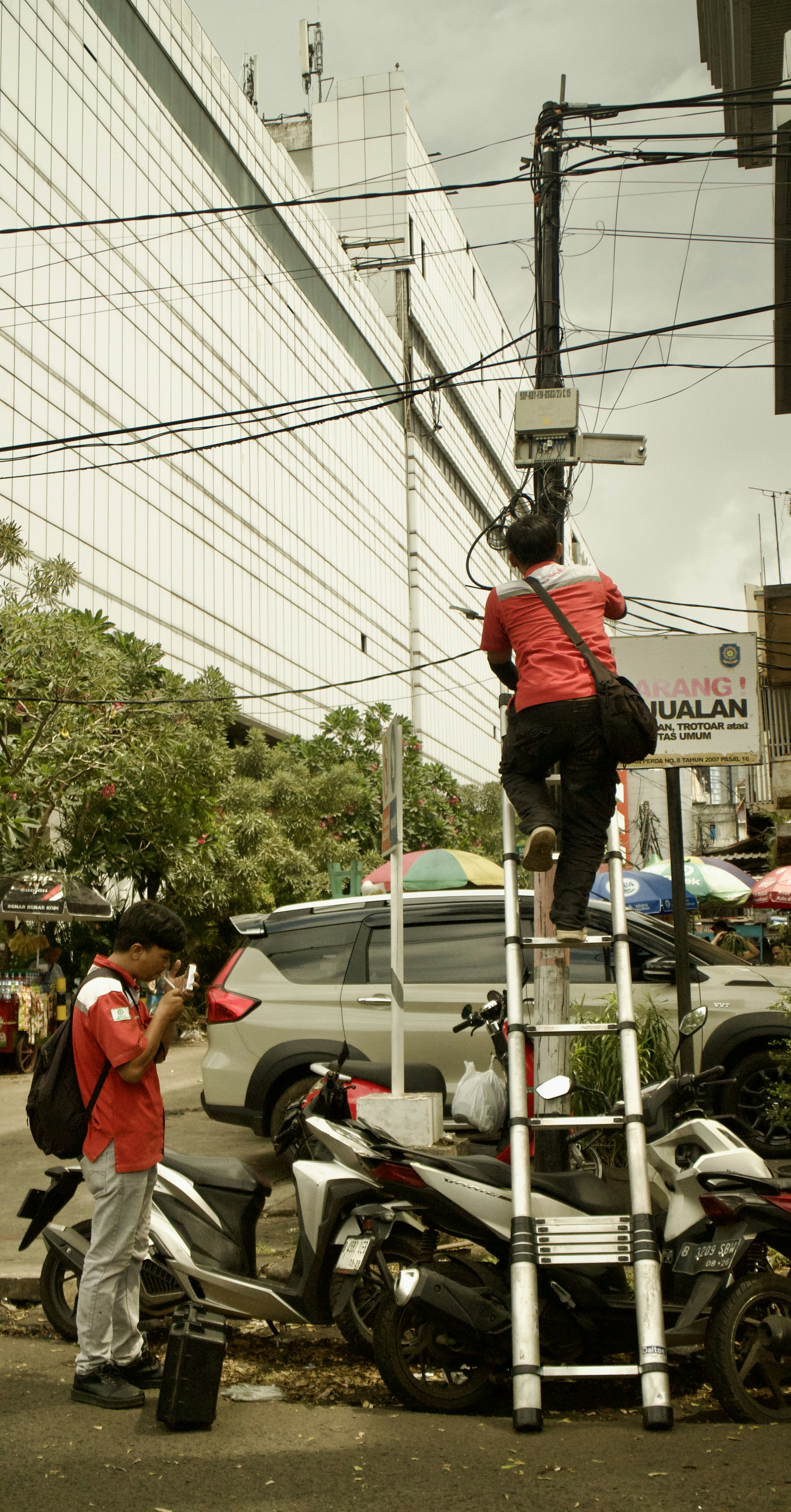 Man on ladder works on wires, another man stands by.