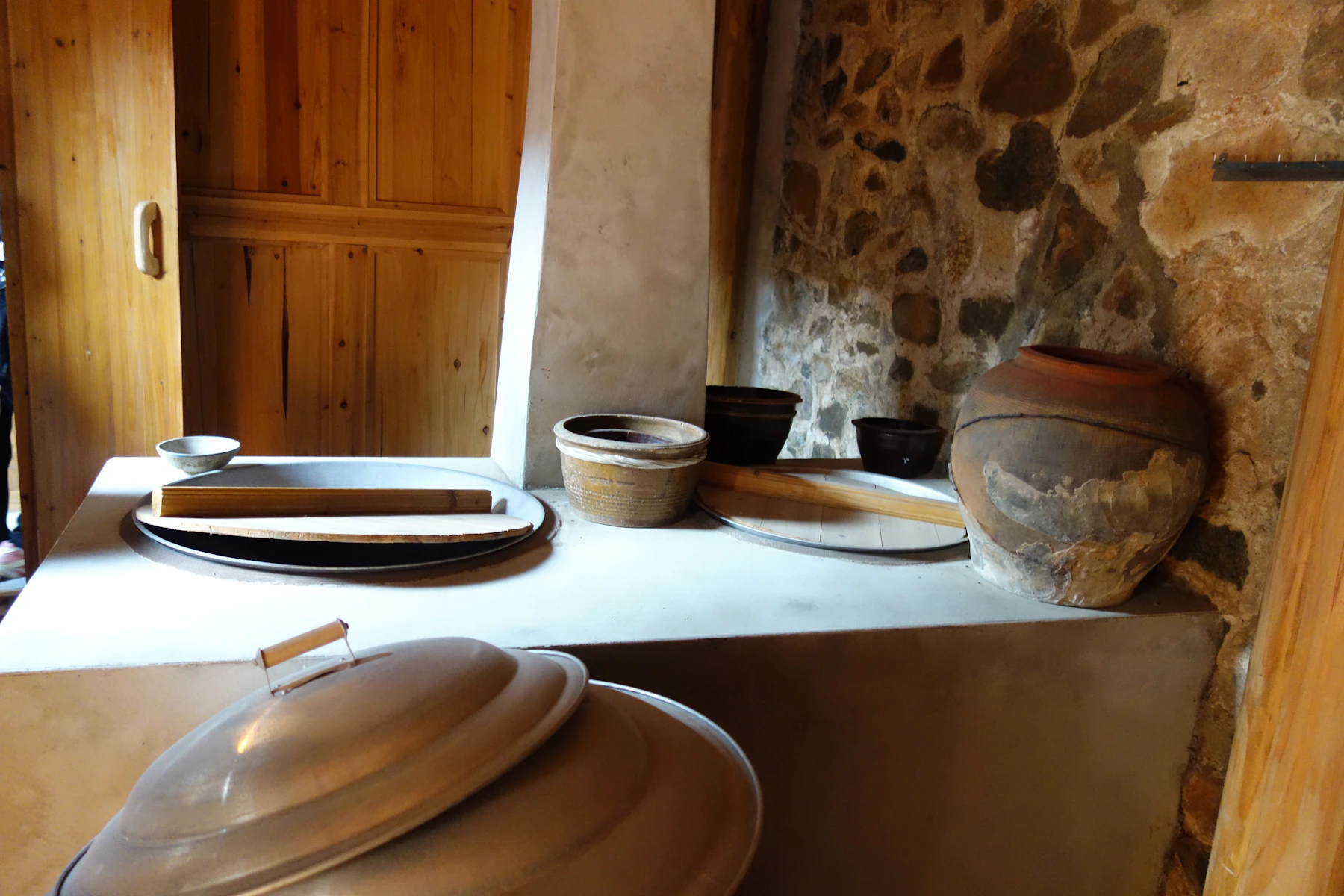Earthenware vessels and wooden trays beside a rough stone wall and wooden door