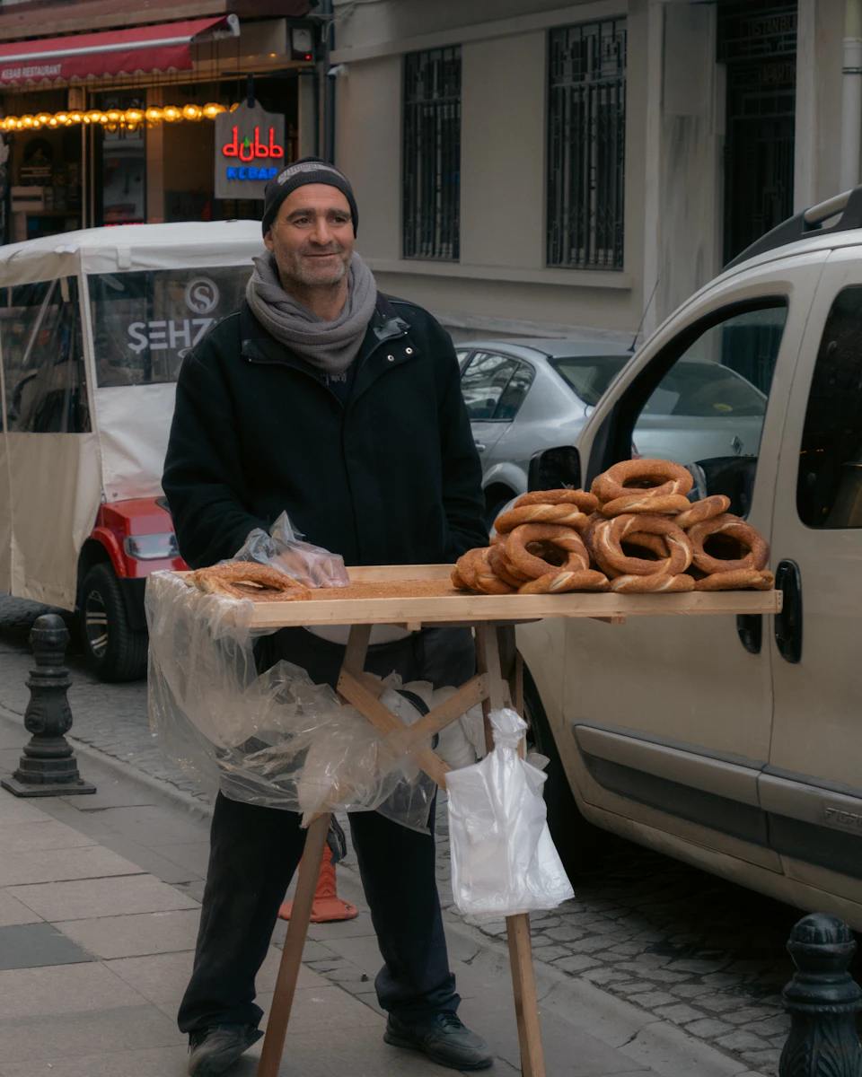 In the image, a man is standing on a sidewalk next to his food cart, which is covered with plastic bags and filled with an assortment of pastries including bagels and donuts. The man is wearing a black jacket and a gray scarf, and he has a warm smile on his face as he looks directly at the camera.

The street scene behind him features parked cars along the side of the road, indicating that this location might be in an urban setting or near a busy commercial area.