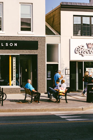 People sitting on benches outside shops on a sunny day.