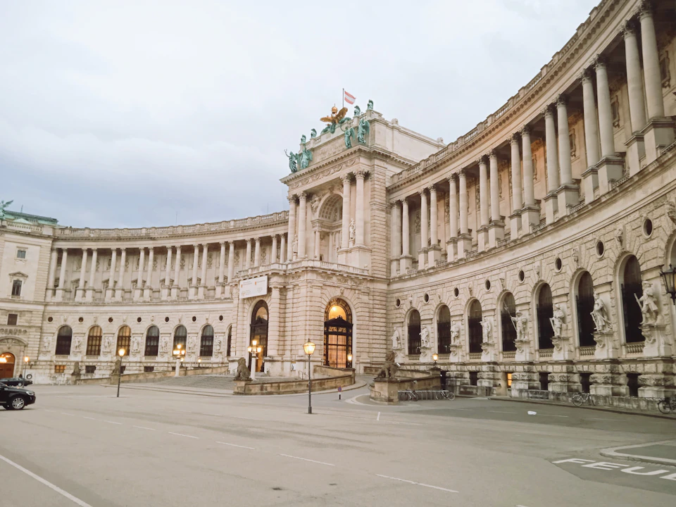 The image shows the Hofburg Palace, a renowned landmark located in Vienna, Austria. The palace is an example of classical architecture and features numerous columns and arches that contribute to its grandeur. The photograph captures the palace from a low angle, emphasizing its imposing stature against the sky. The perspective gives a sense of looking up at the palace's impressive height and architectural details.