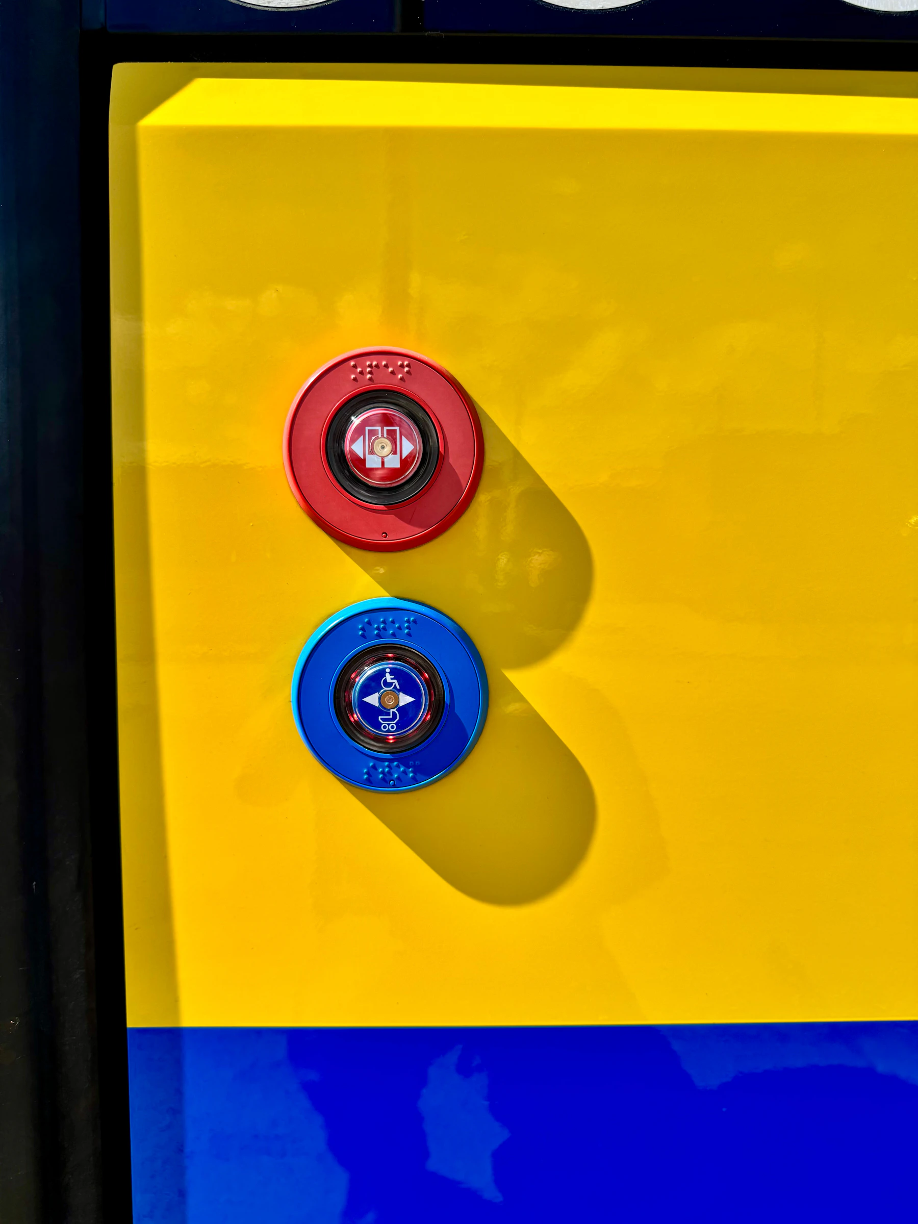 A red and a blue door button on a yellow panel, each ringed with raised braille dots