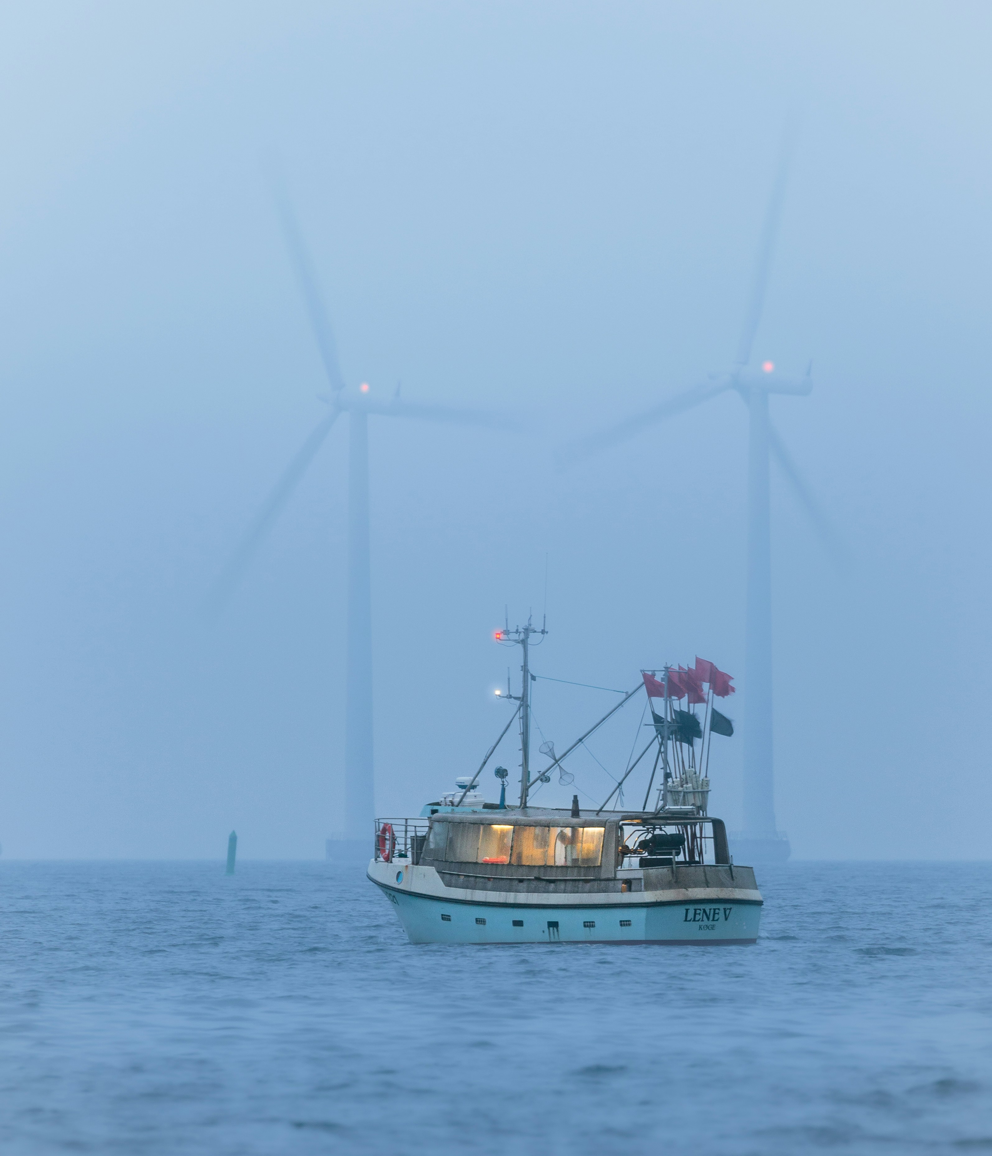 Fishing boat with its deck lights on and marker flags flying, idling in thick fog with wind turbines faint behind