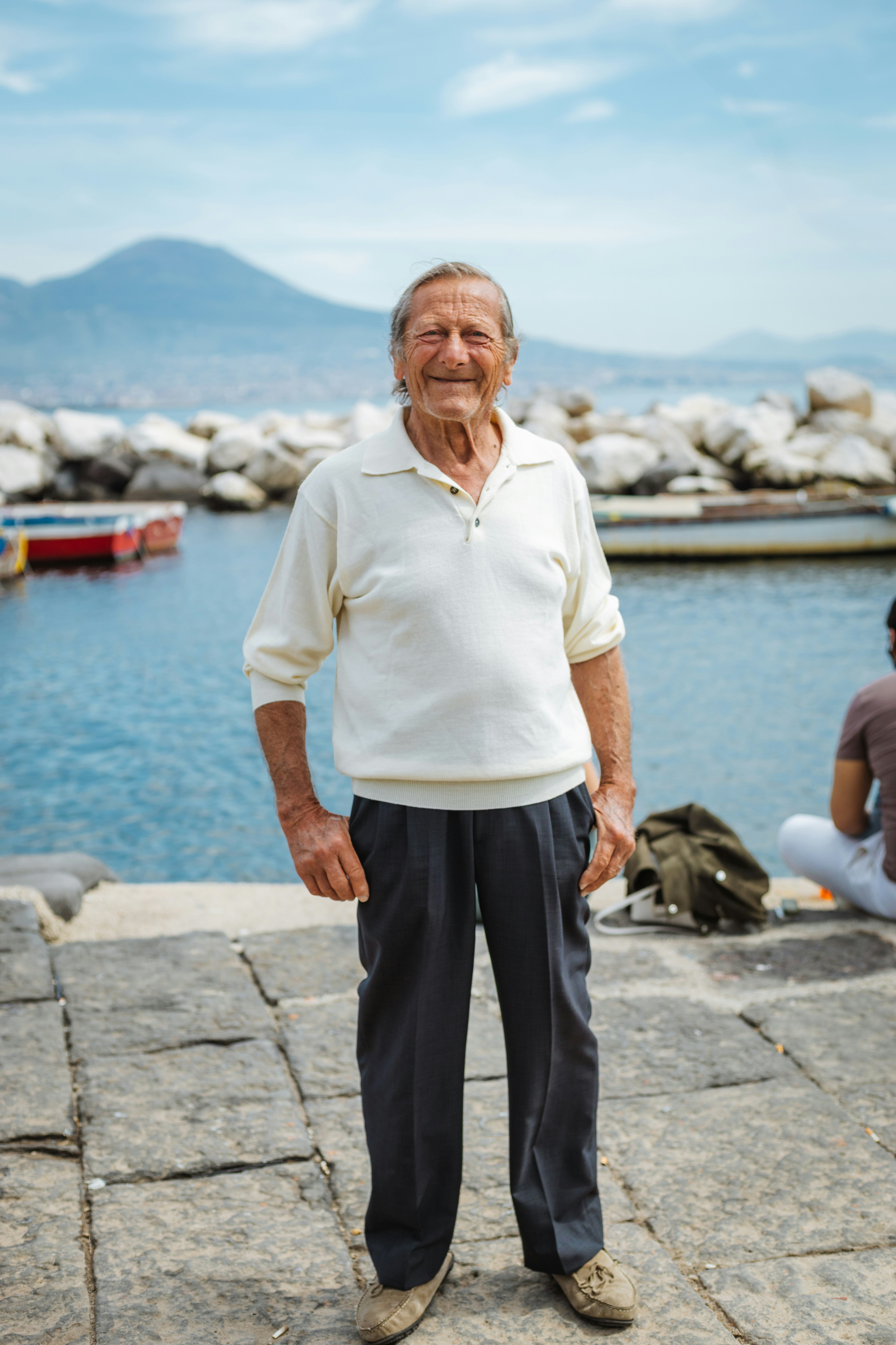 Elderly man standing by the water with a mountain background.