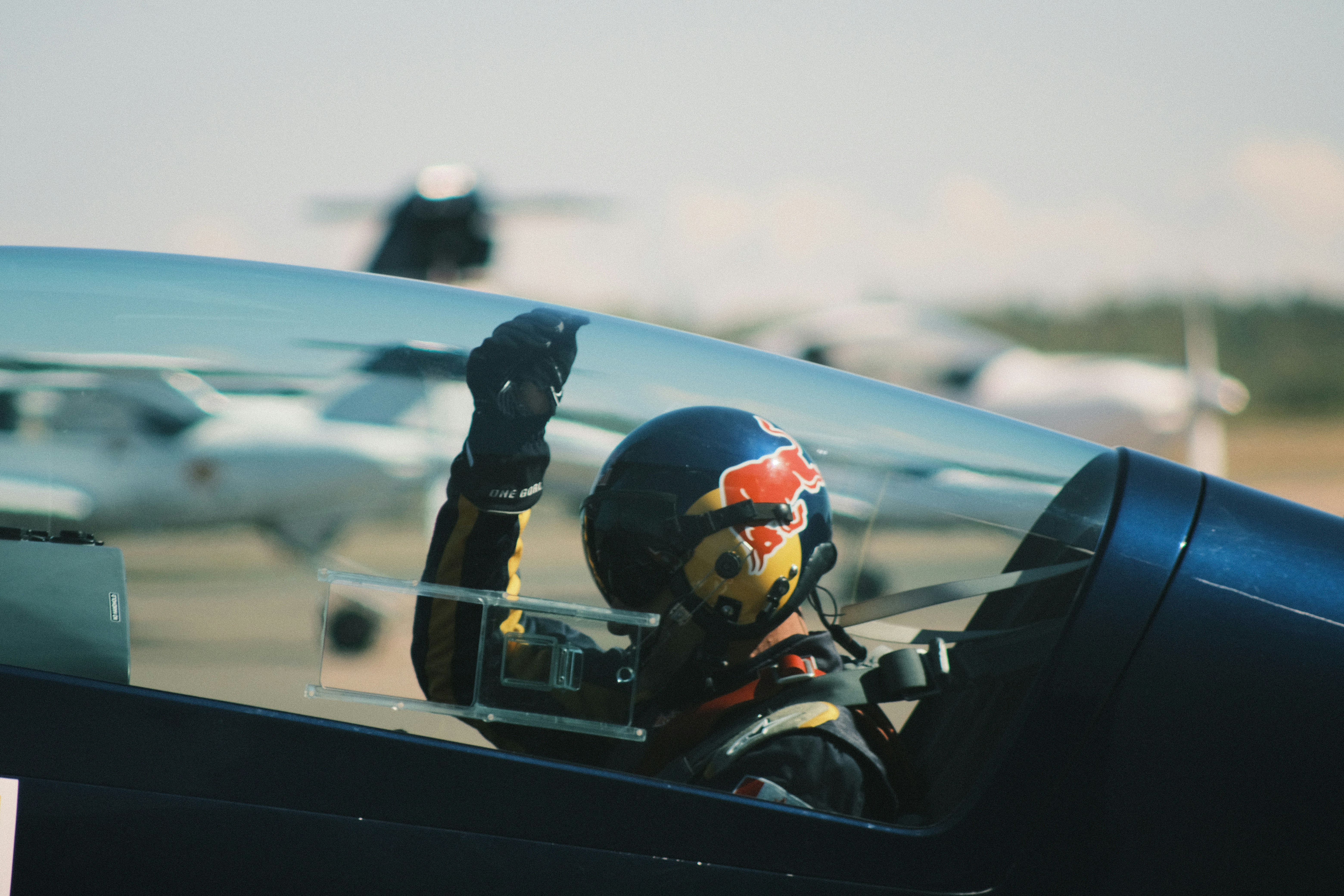 Pilot in cockpit waves with red bull helmet.