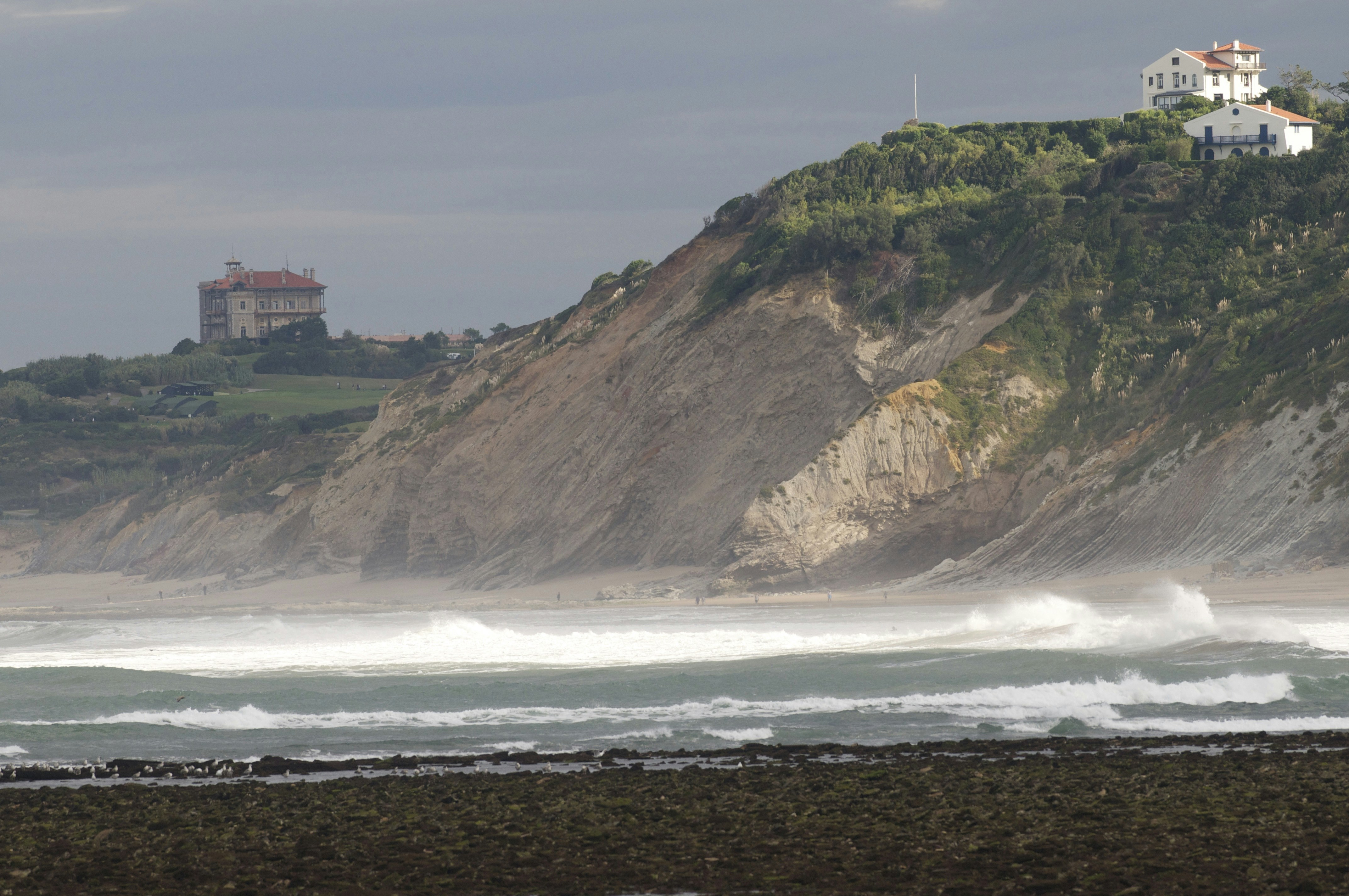 Waves crash on a rocky shore below coastal buildings.