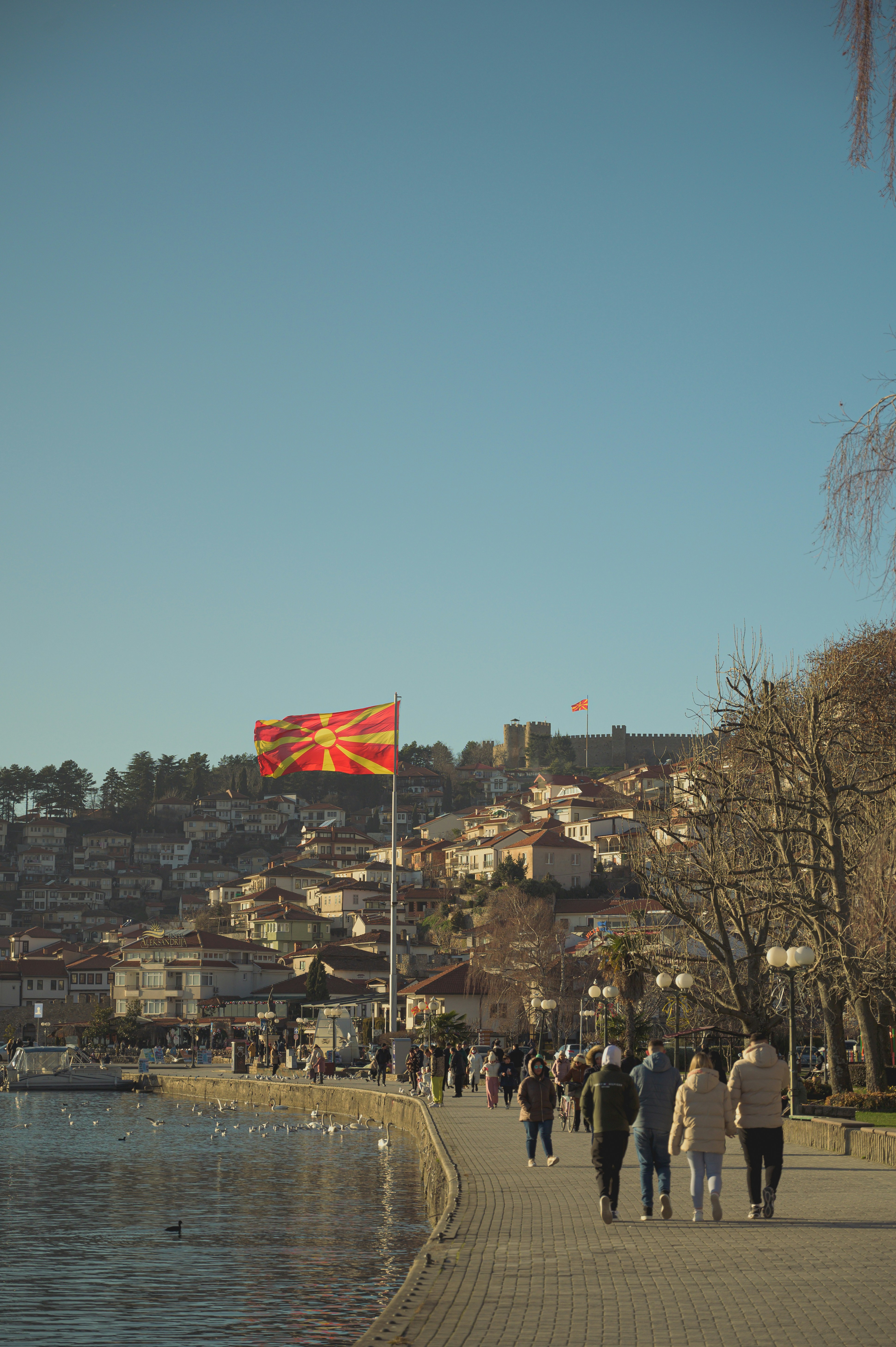 People stroll along a waterfront promenade with a flag.