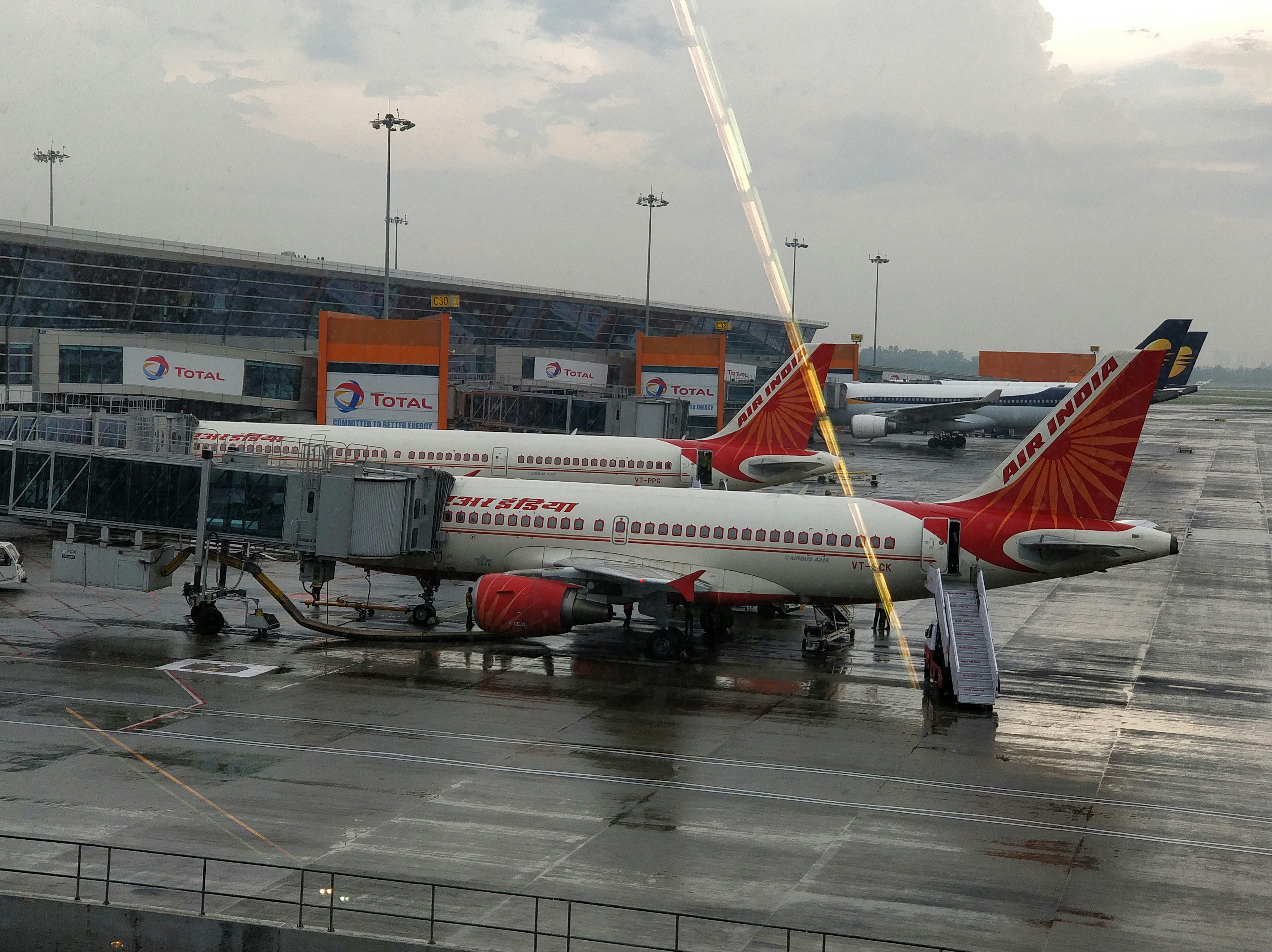Air India विमान एक हवाई अड्डे पर खड़ा है, जिसके पीछे एक डिजिटल बोर्ड पर 