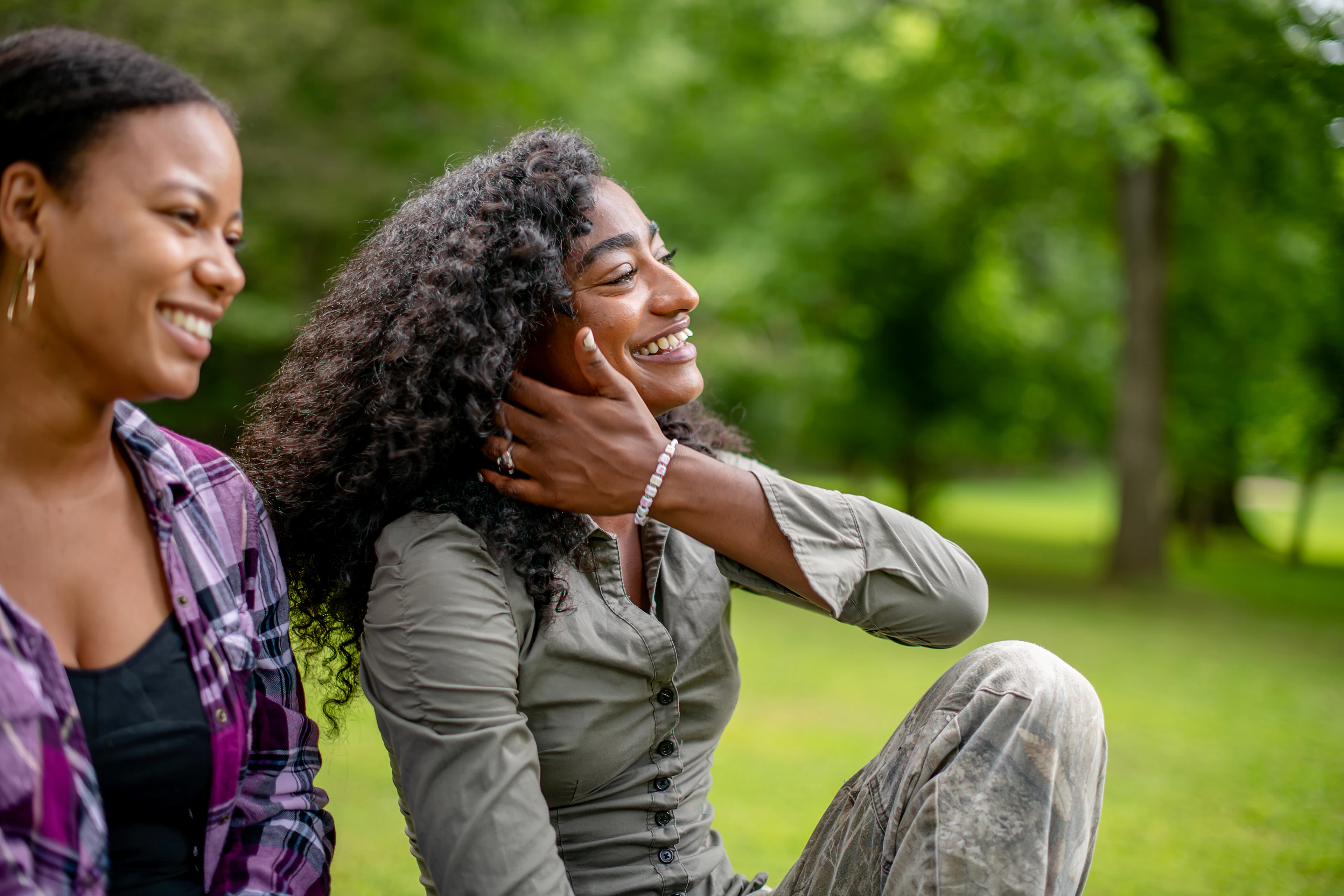 A woman in an olive top laughing in a green park, her hand at her cheek
