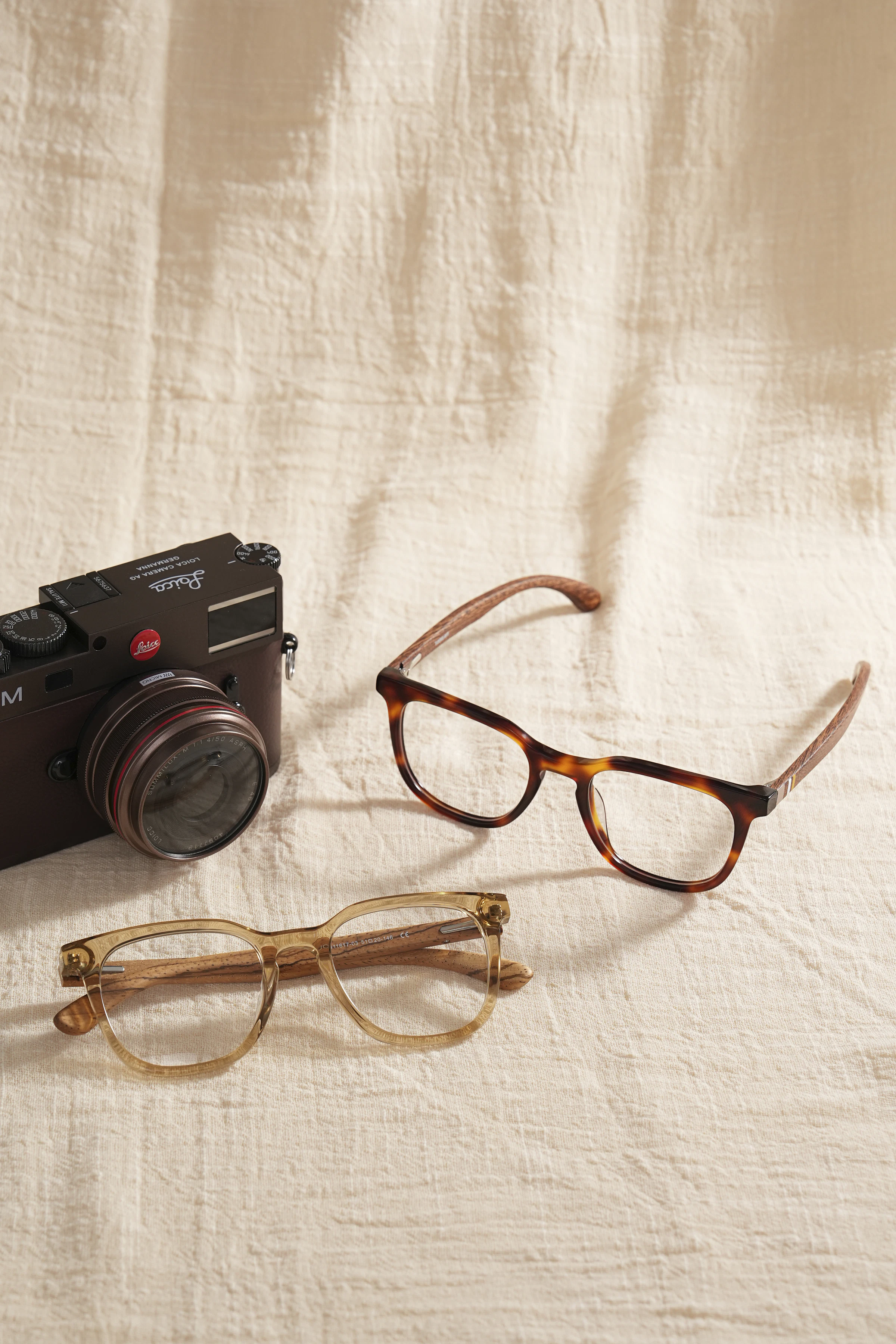 Deux paires de lunettes acétate posées sur lin écru, à côté d'un appareil Leica