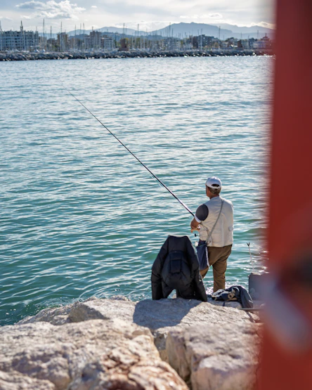 Man fishing from rocky shore with city in background