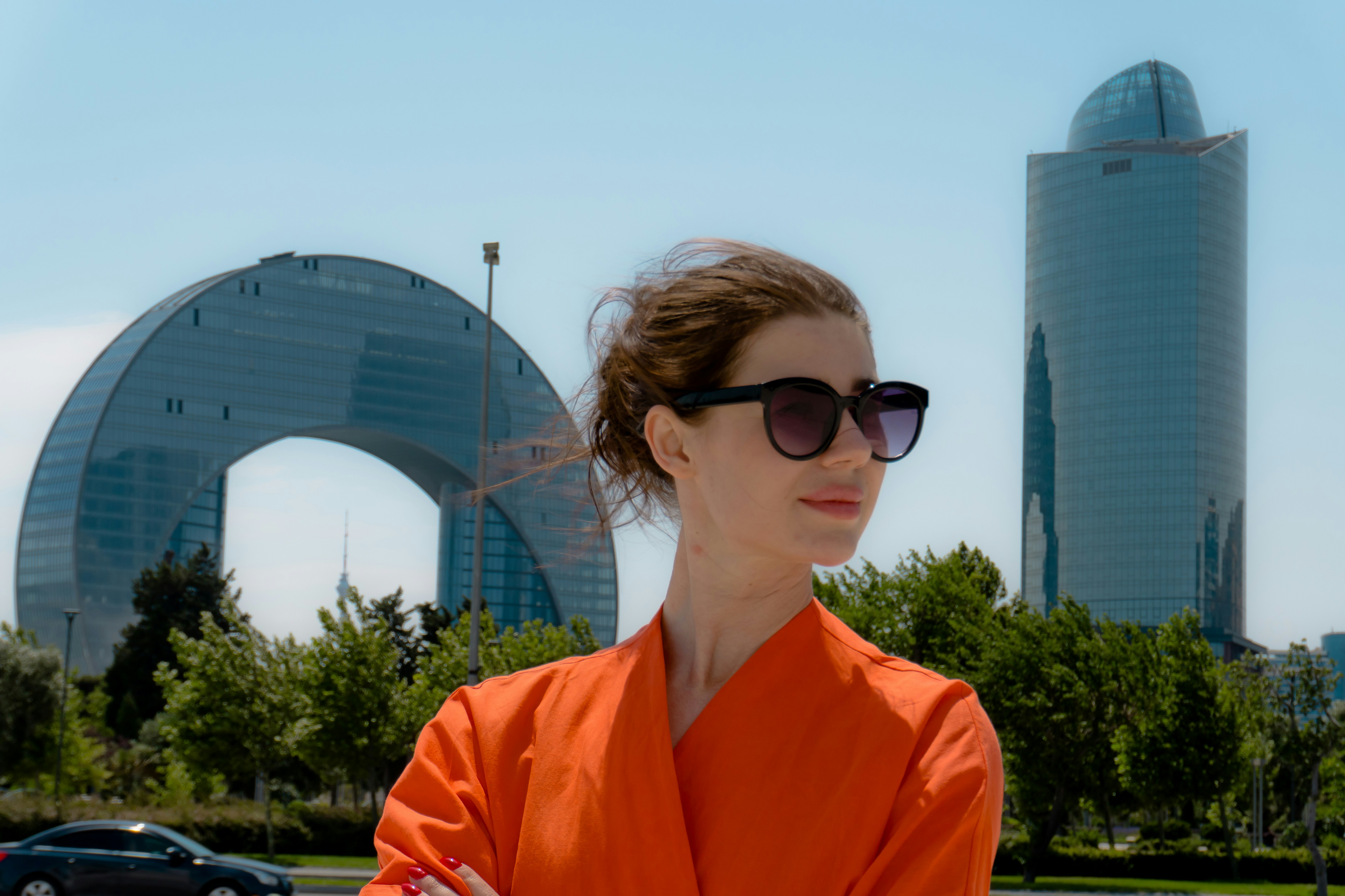 Woman in orange with sunglasses, modern buildings behind.