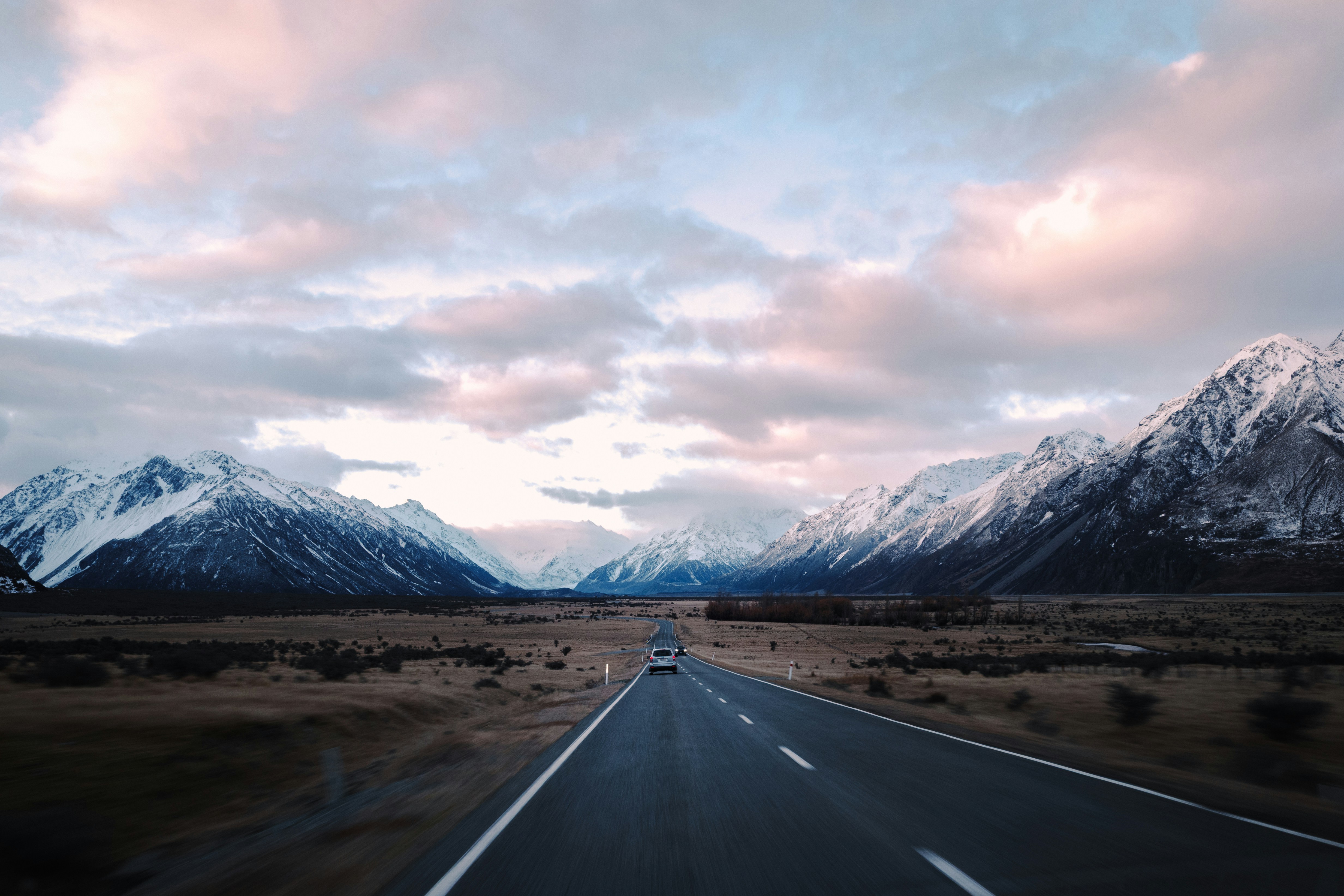 A car drives down a road between snow-capped mountains.