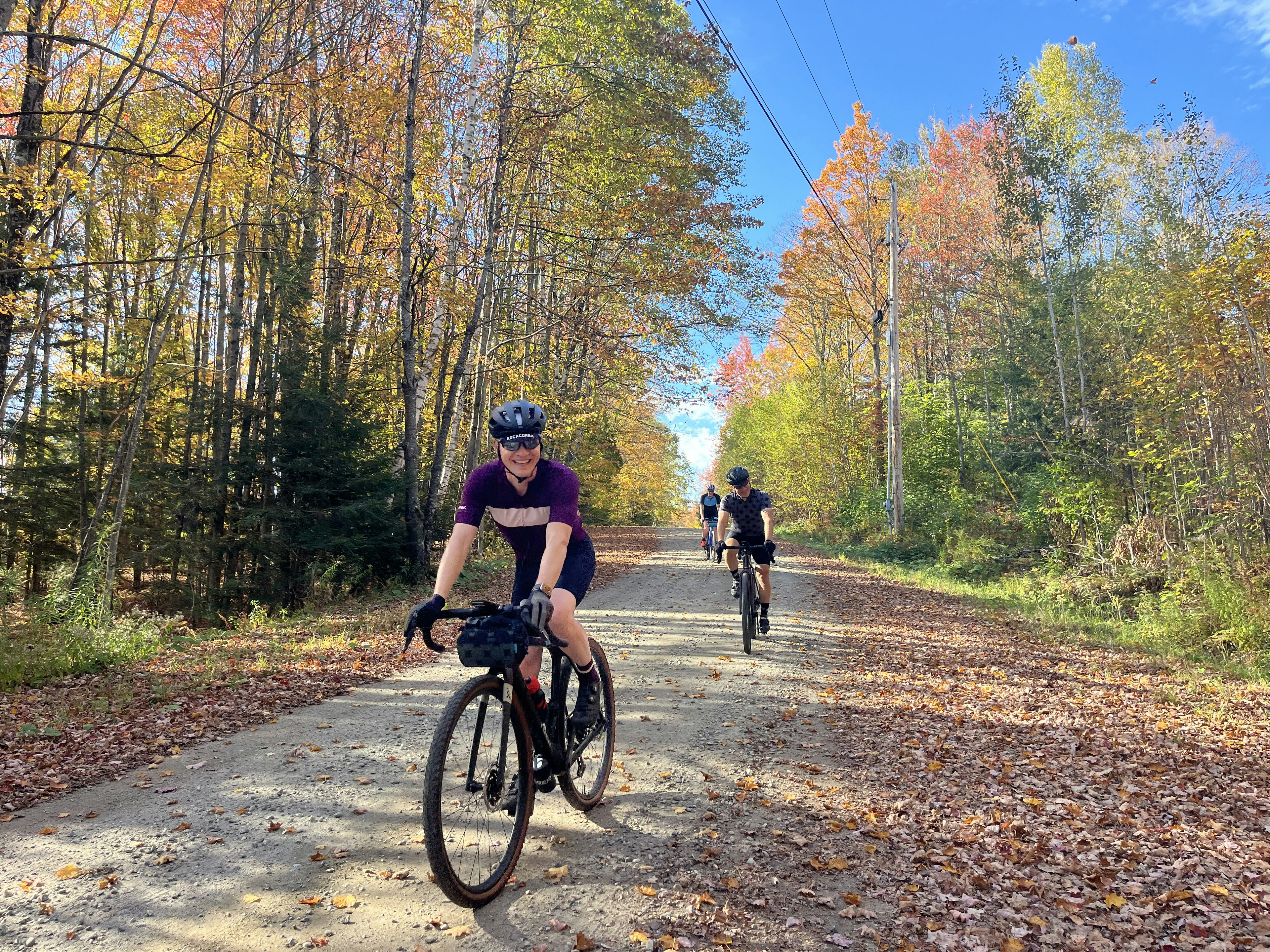 Cyclists ride on a gravel path surrounded by autumn trees.