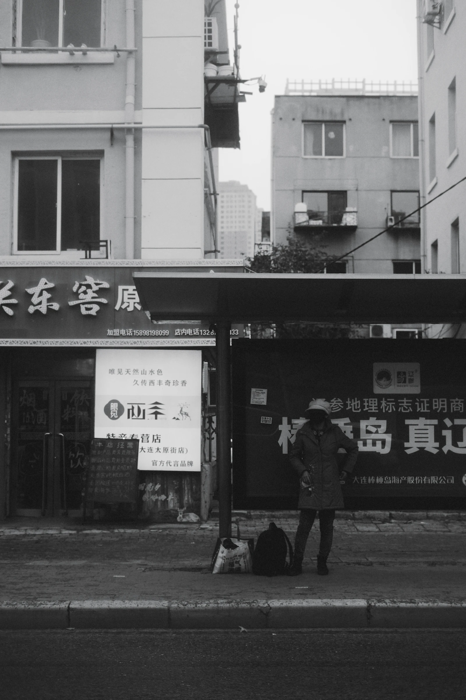 A traveller standing at a bus stop with a backpack and shopping bags.