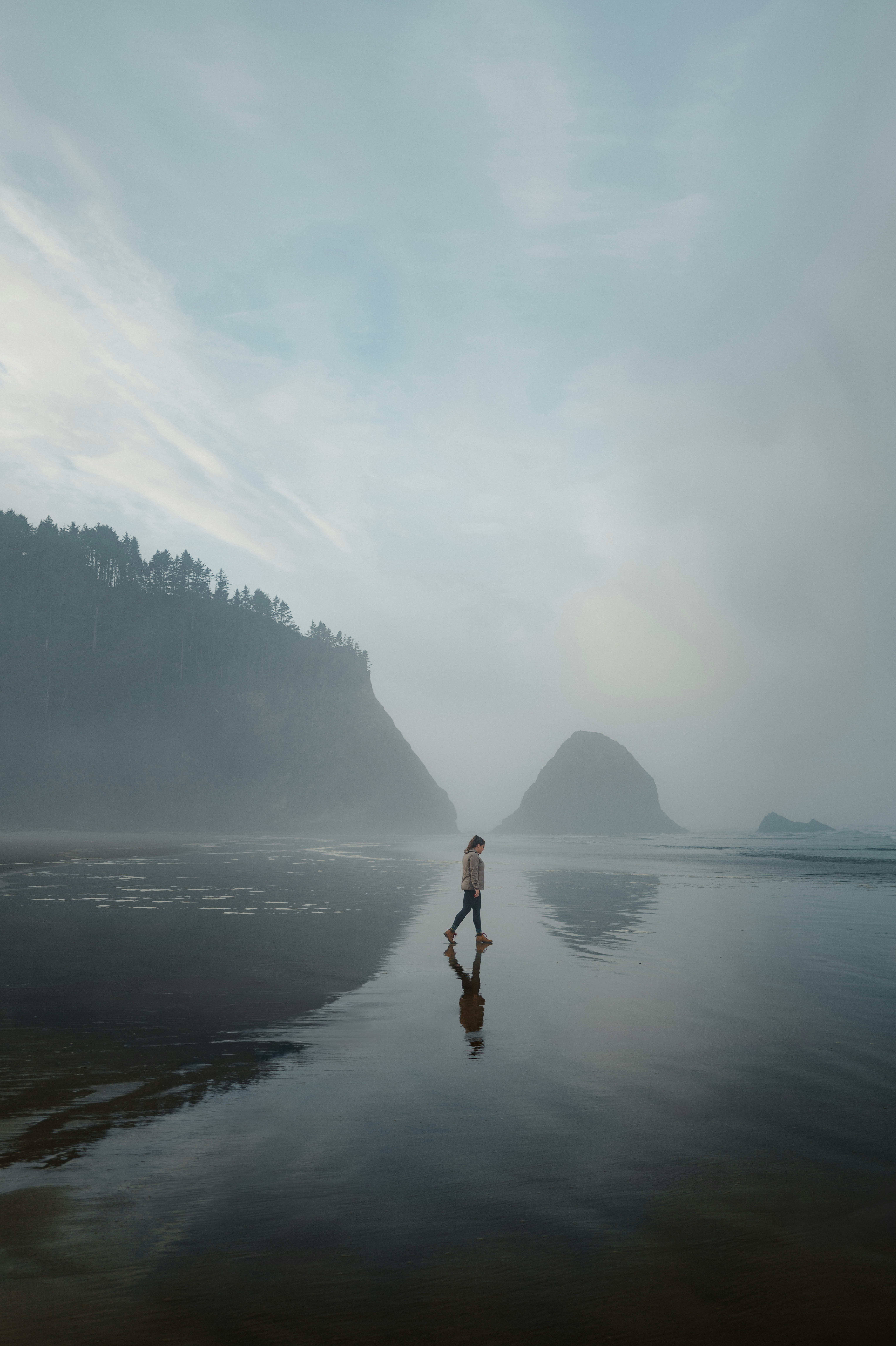 Una persona cammina su una spiaggia nebbiosa con riflessi.