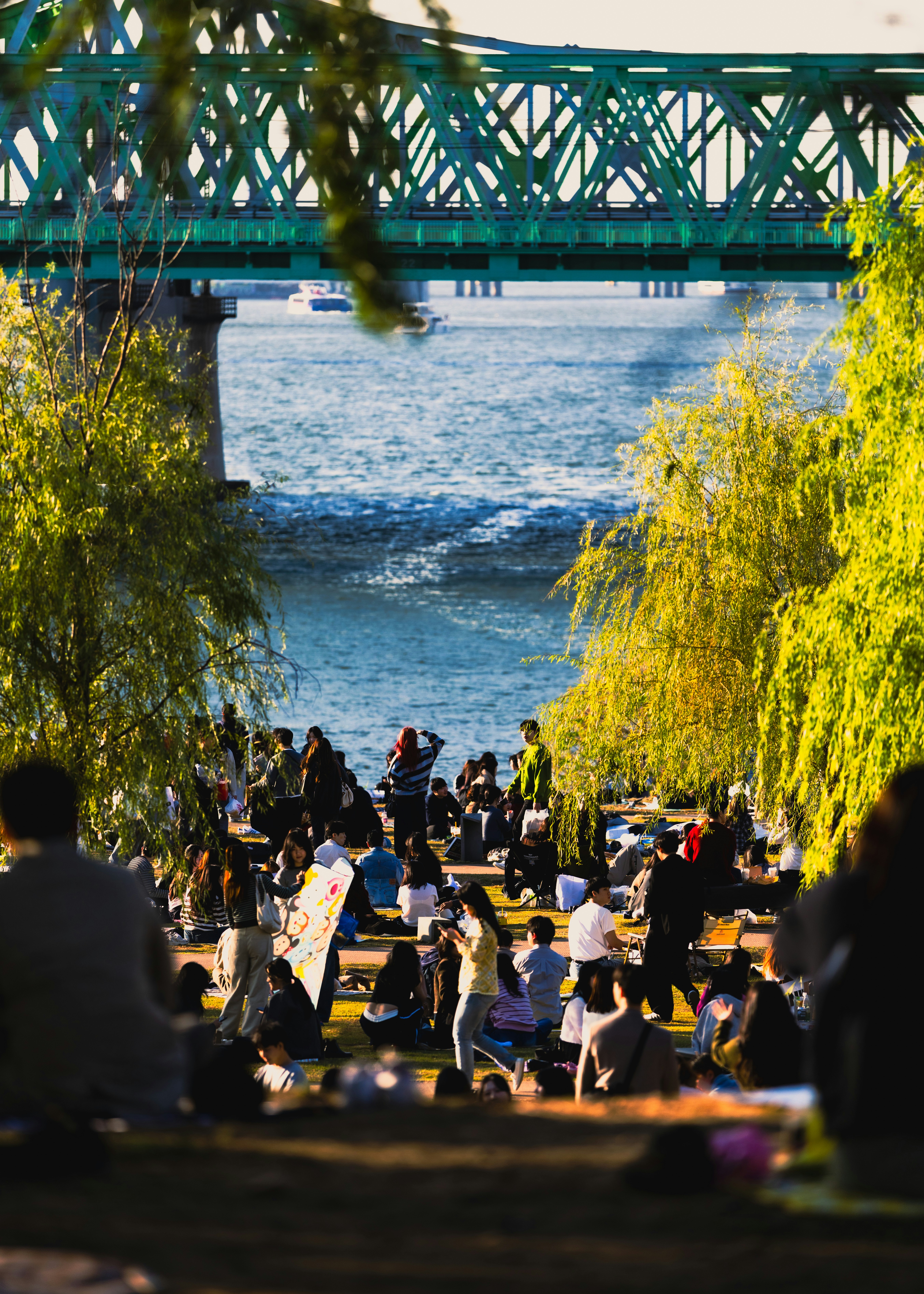 People relaxing by a river under a green bridge.
