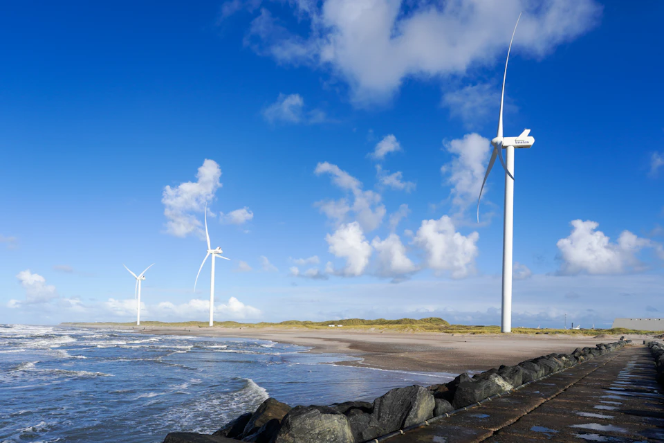 Three wind turbines stand on a beach with white structures contrasting against blue sky