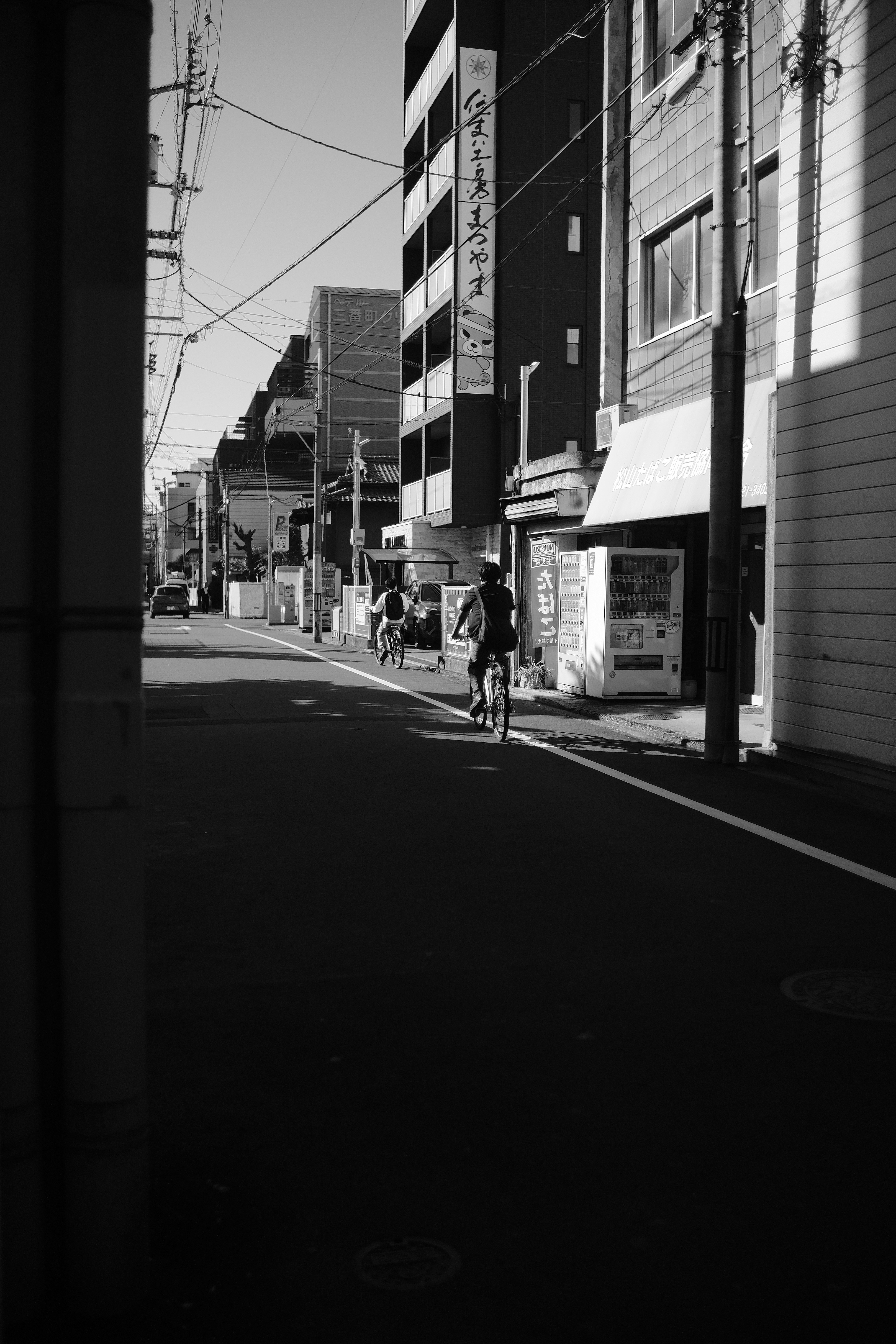 Two cyclists ride down a sunlit street in japan.