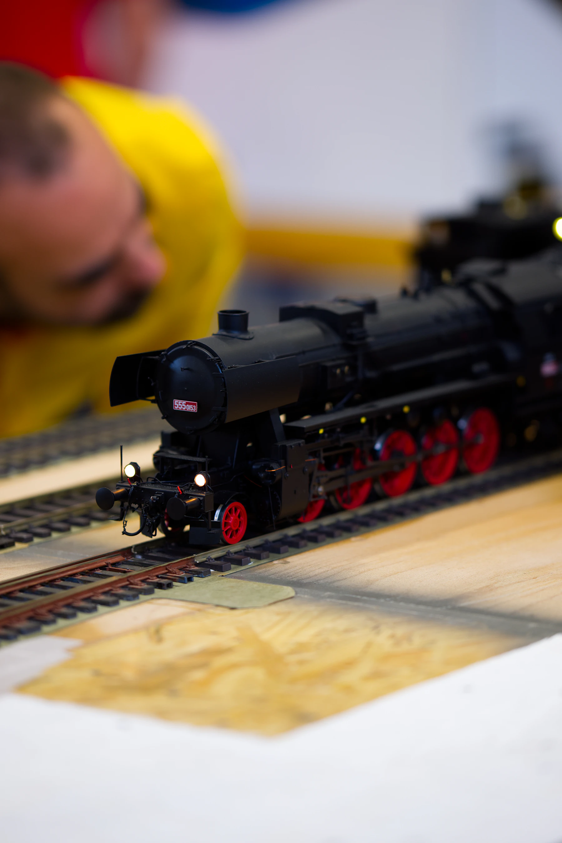 A black model steam engine with red wheels on track laid over bare plywood, with a man leaning in to look at it