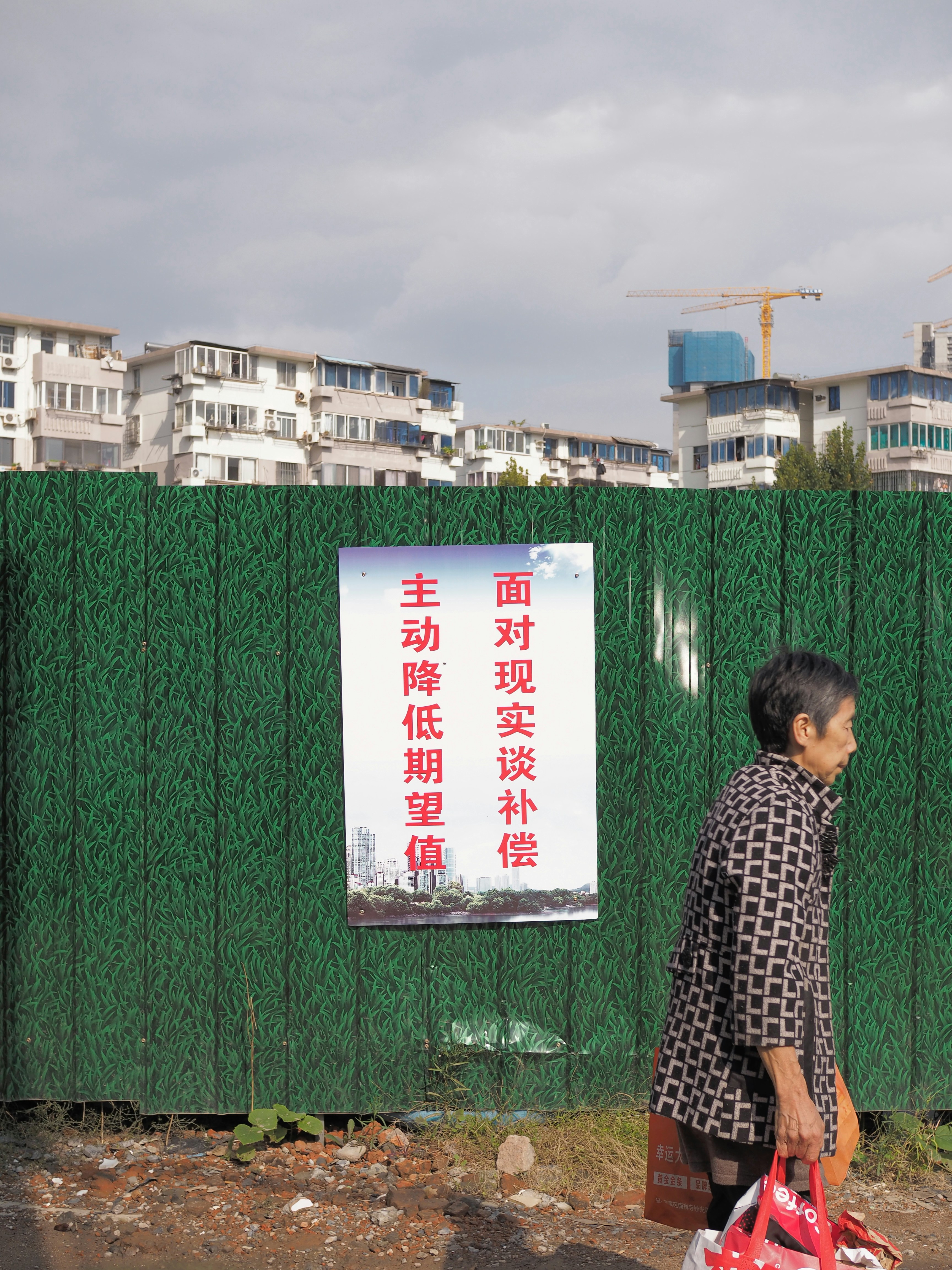 Elderly woman walks past construction site fence with chinese sign.