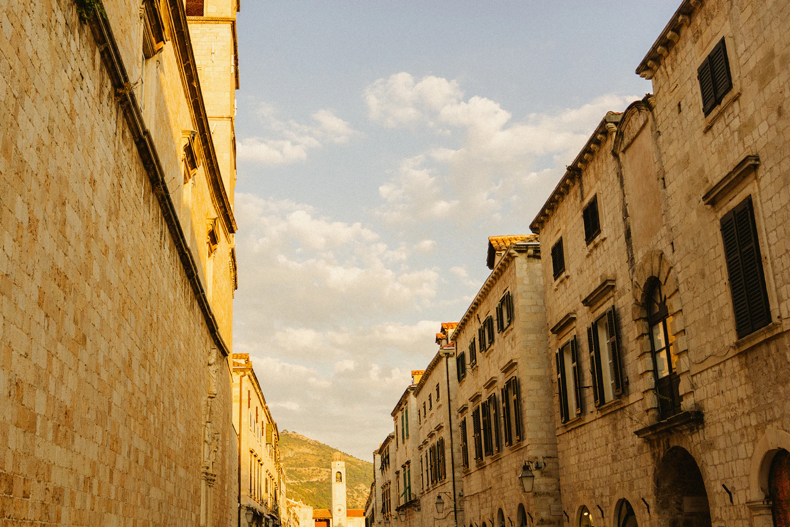 A narrow limestone side street in Dubrovnik's Old Town with the city bell tower and Mount Srd in the distance