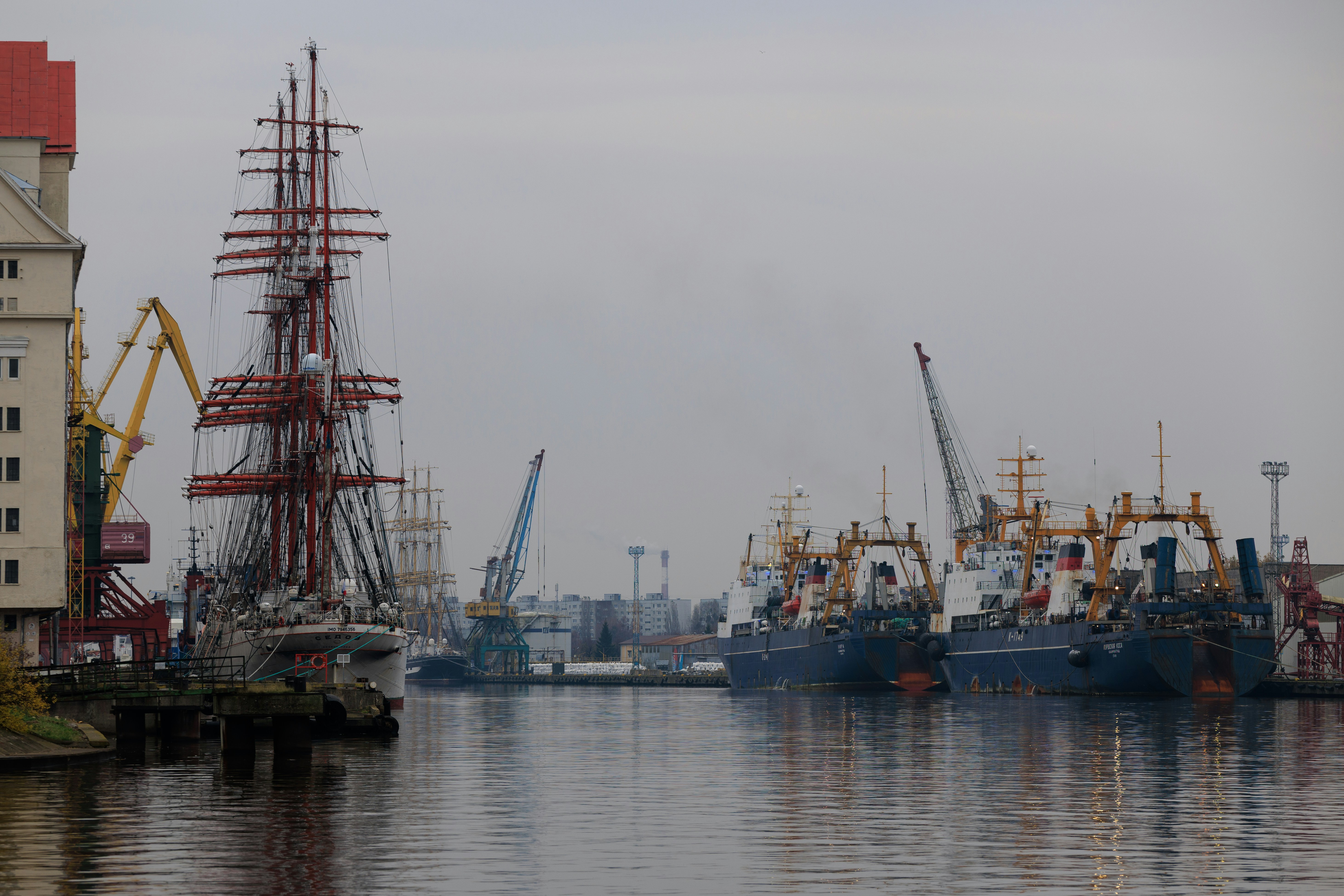 The four-masted barque Kruzenshtern stands with its tall rigging next to working fishing boats and cranes at the Pregolya River port in Kaliningrad on a grey day.