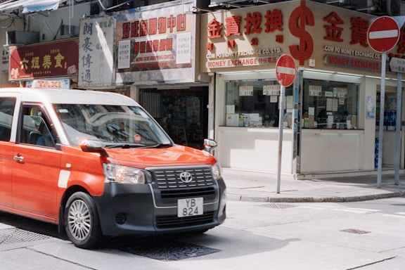 Red taxi drives past shops on city street.