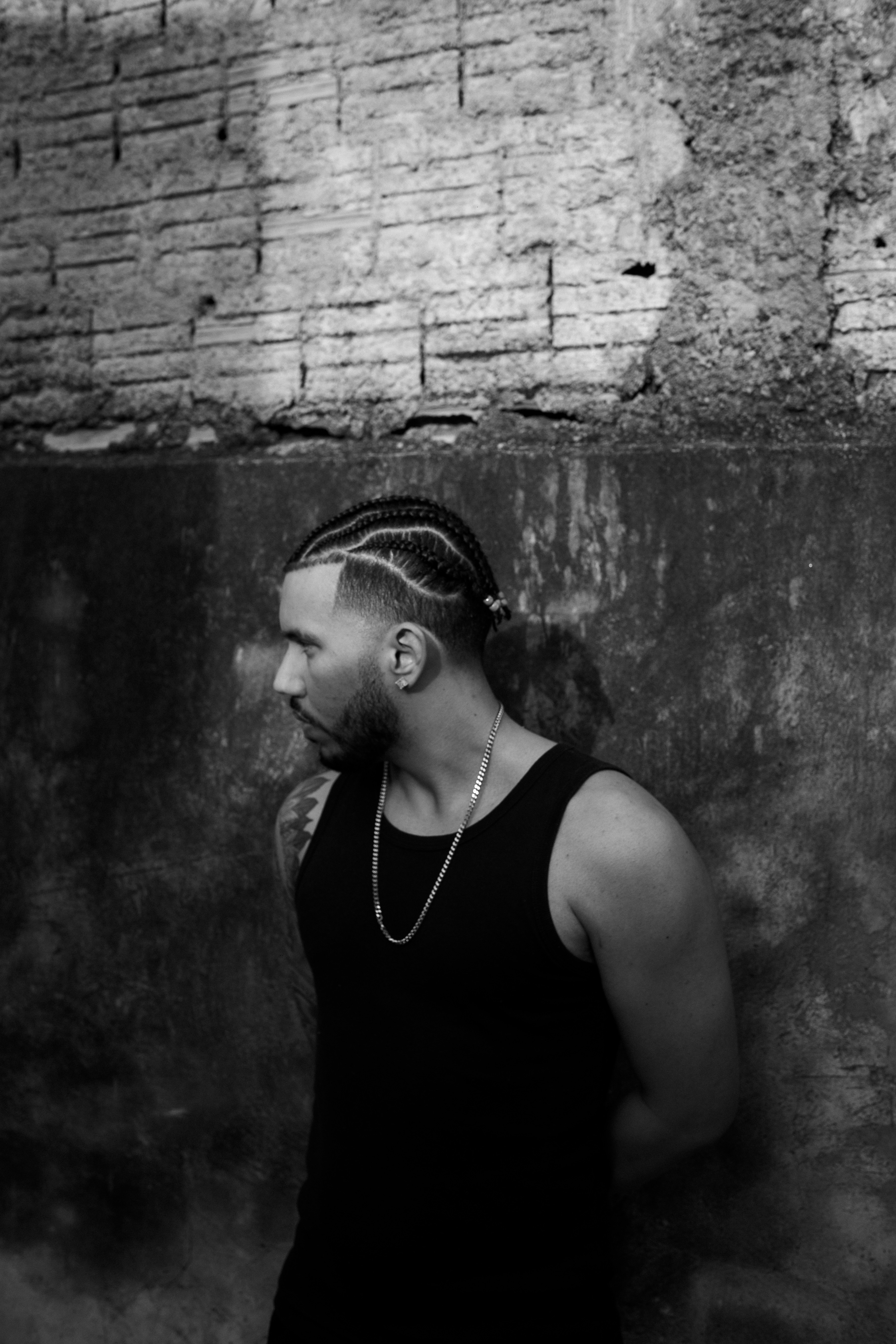 Man with braided hair leans against a textured wall.