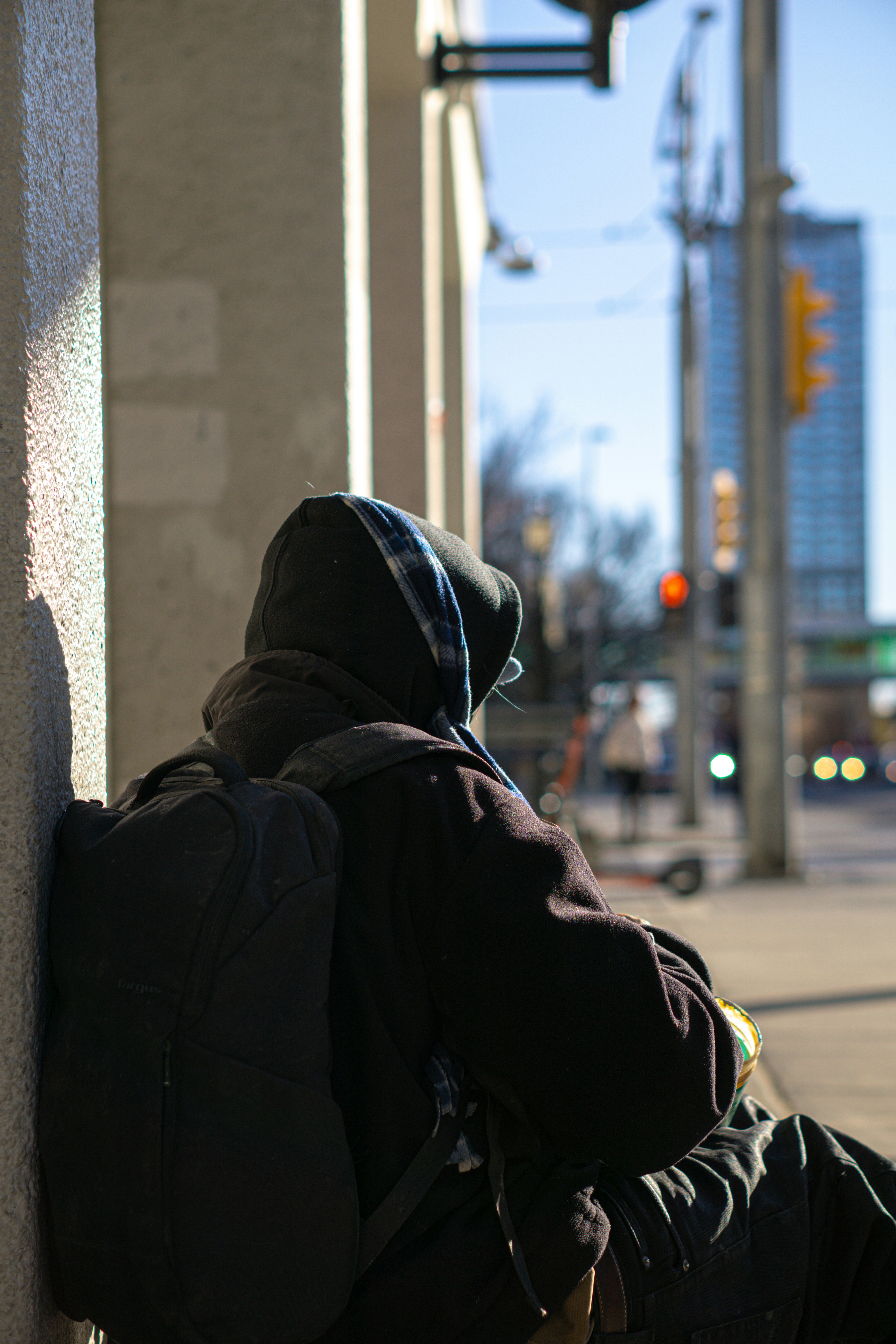 Person in hoodie with backpack sits by wall outdoors.