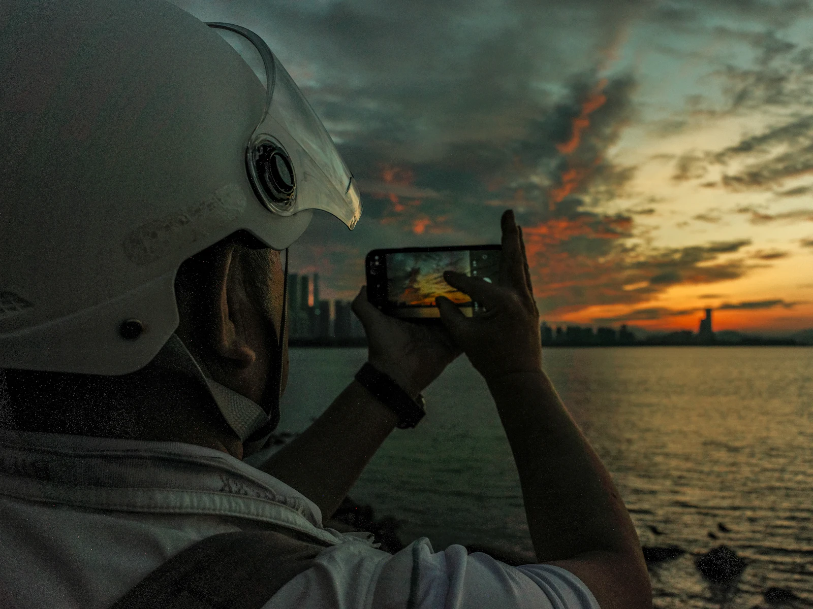 Traveller photographing a dramatic orange sunset over a city skyline and waterfront with a smartphone