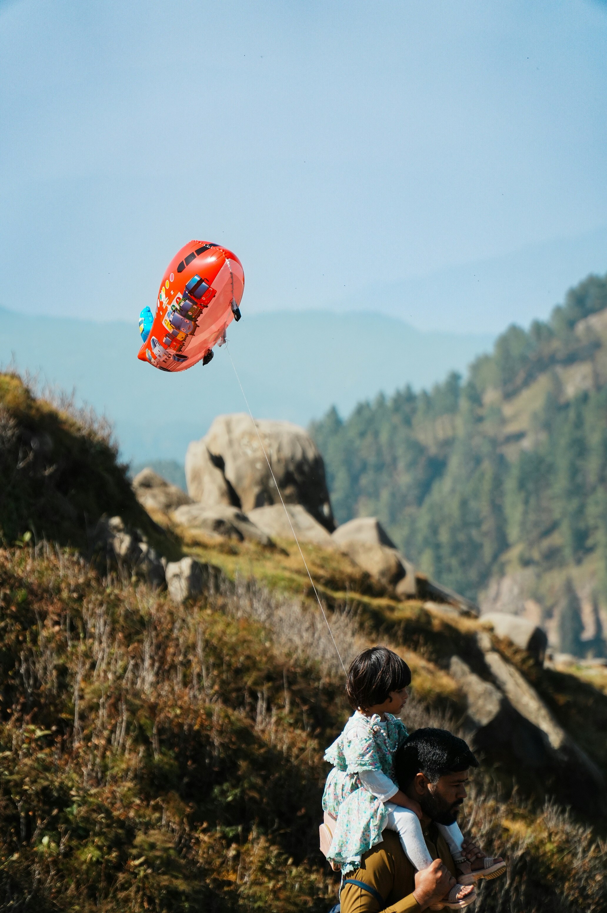 Father carries child with balloon in mountainous landscape
