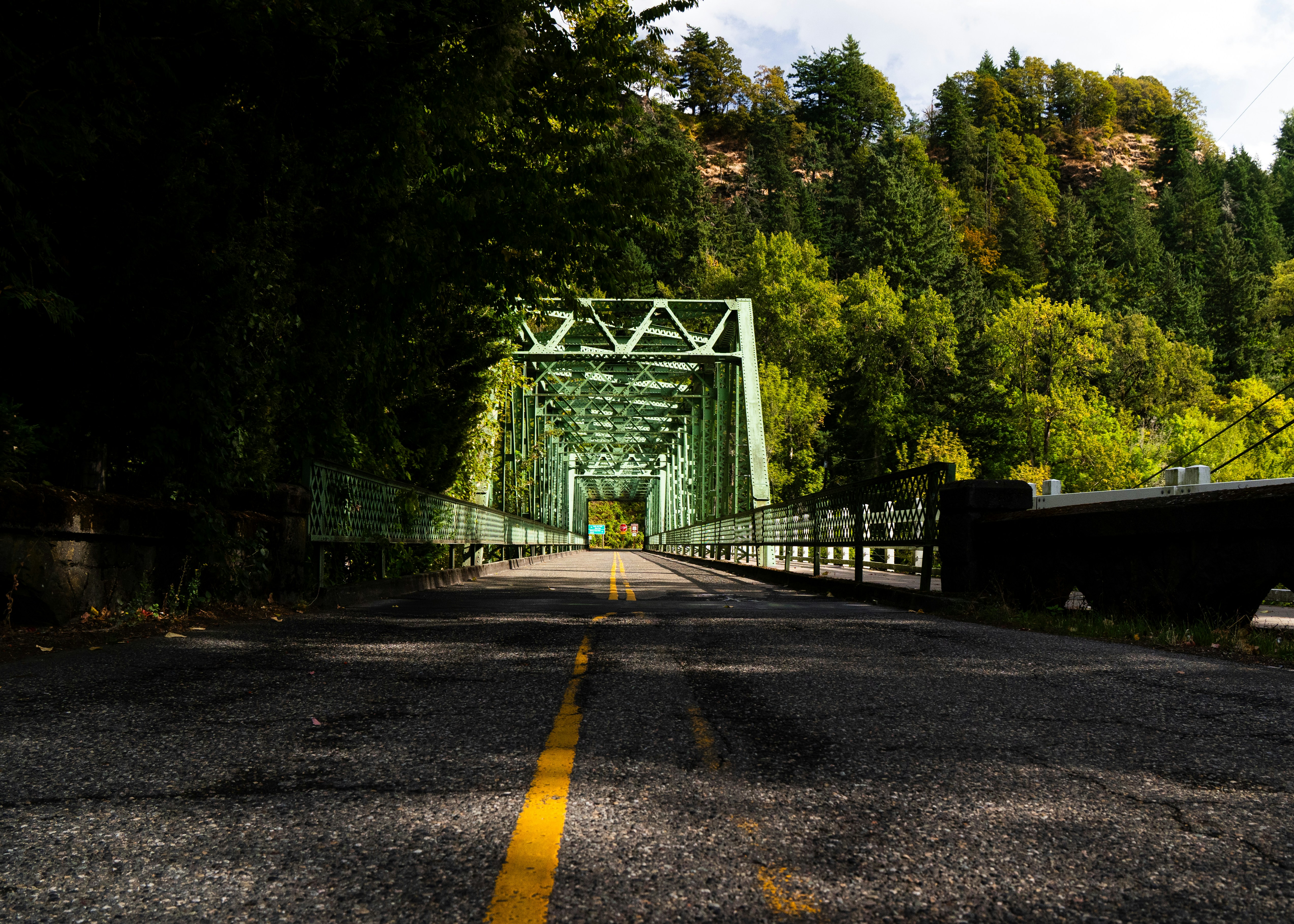 Green truss bridge over a road surrounded by trees.