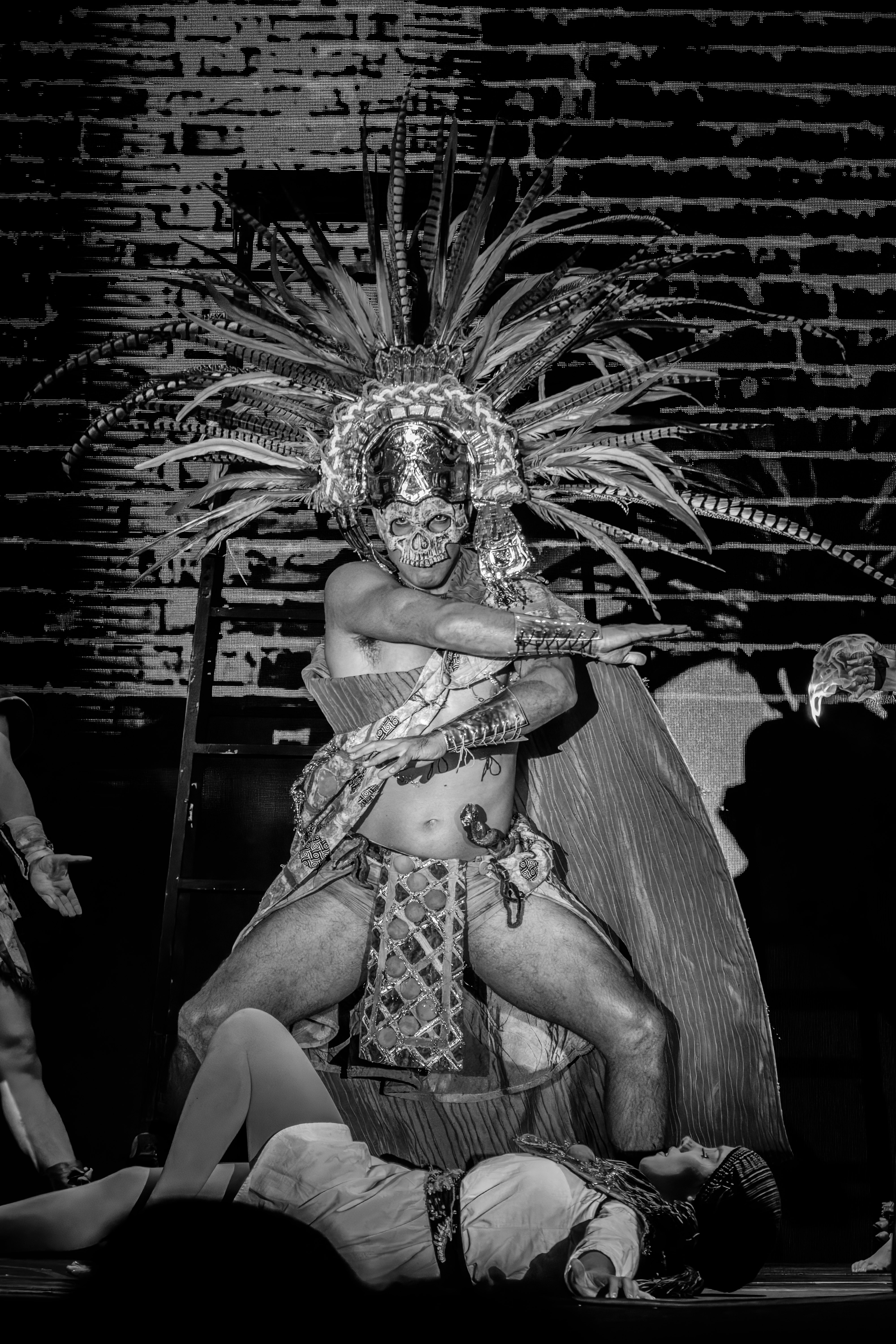 Man in elaborate headdress and costume over fallen figure.