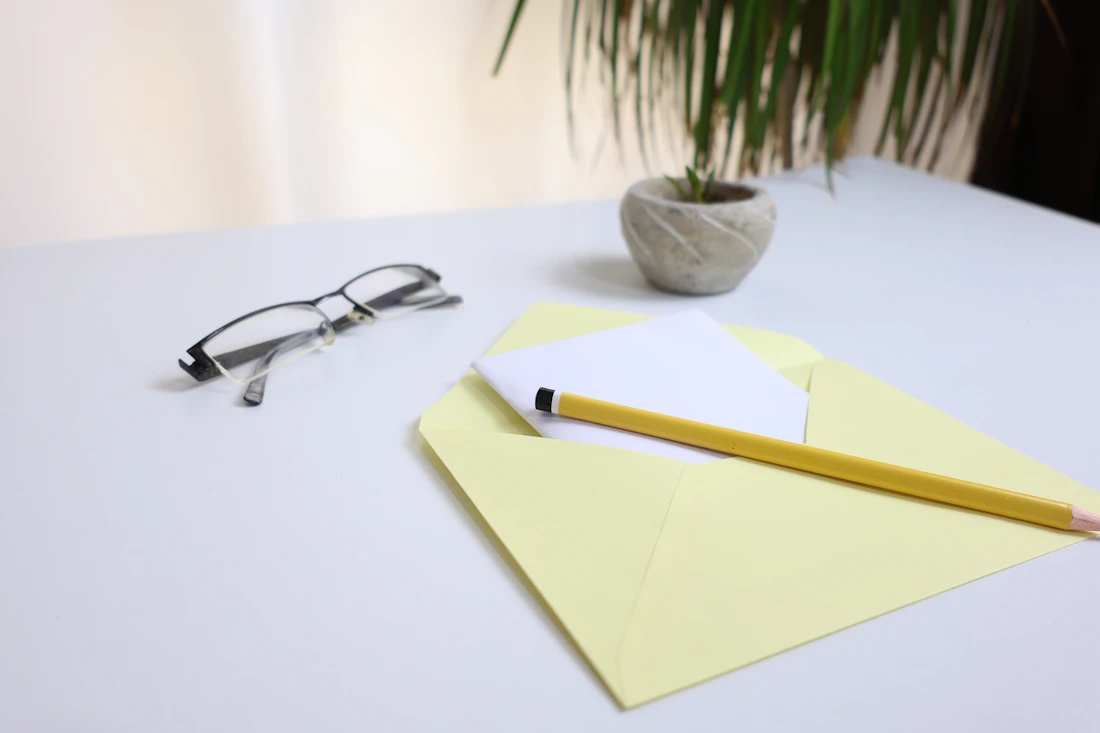 Heller Schreibtisch mit gelbem Umschlag, Stift, Brille und Grünpflanze
