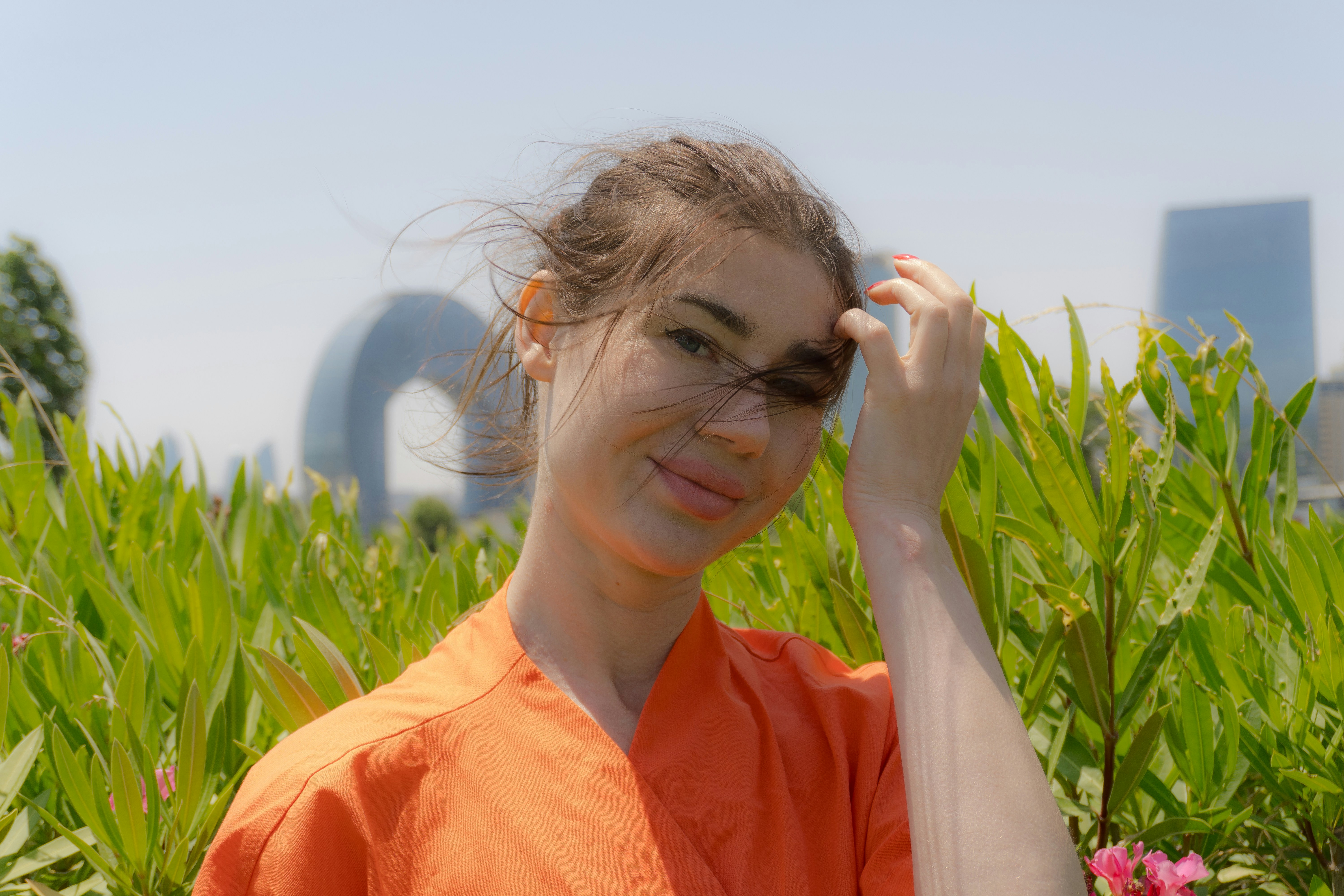 Young woman in orange shirt with greenery and city buildings.