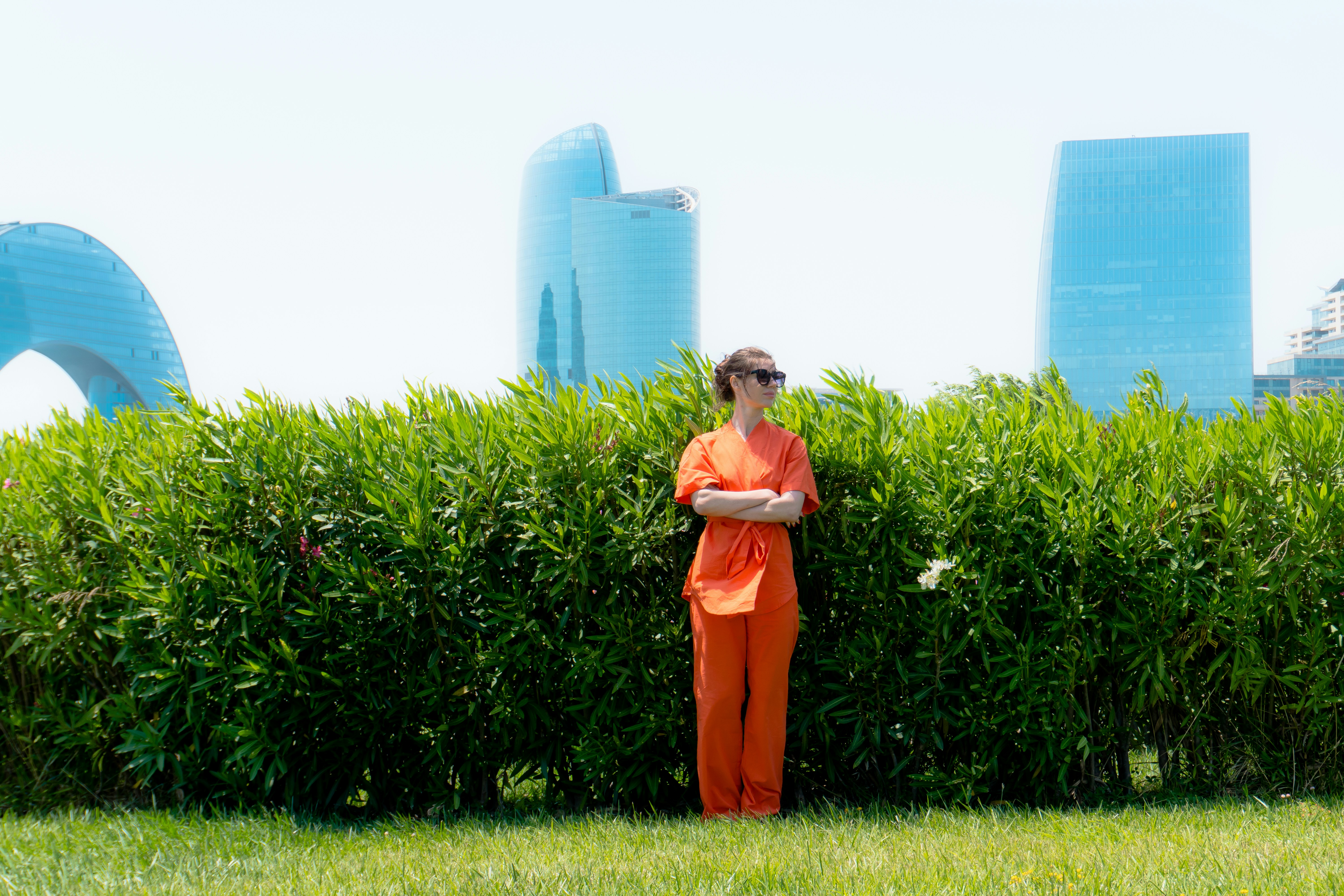 Woman in orange standing behind green hedge with buildings.