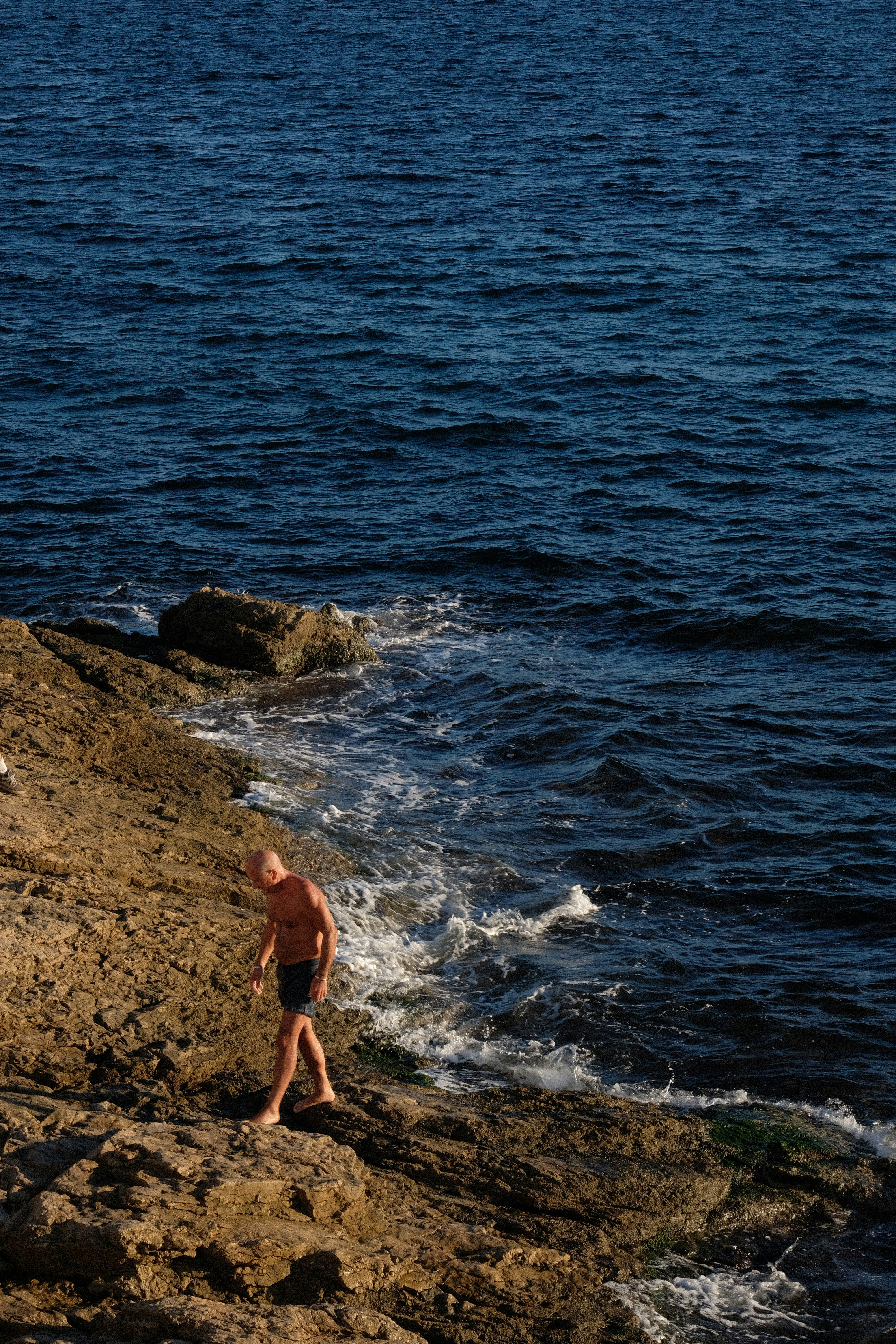 Man walking on rocky shore near dark blue ocean.